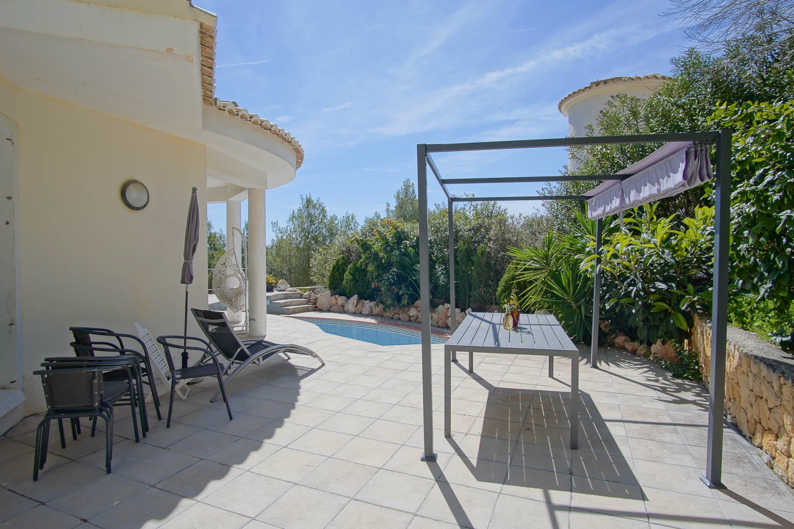 Terrasse extérieure carrelée avec pergola métallique, coin repas et piscine privée entourée d'une végétation méditerranéenne luxuriante.