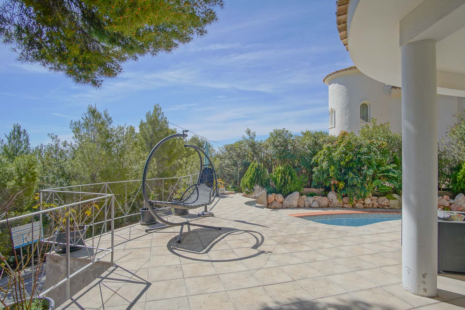 Terrasse extérieure dallée avec fauteuil suspendu, offrant un accès à la piscine et une vue sur la végétation environnante et l'architecture locale.