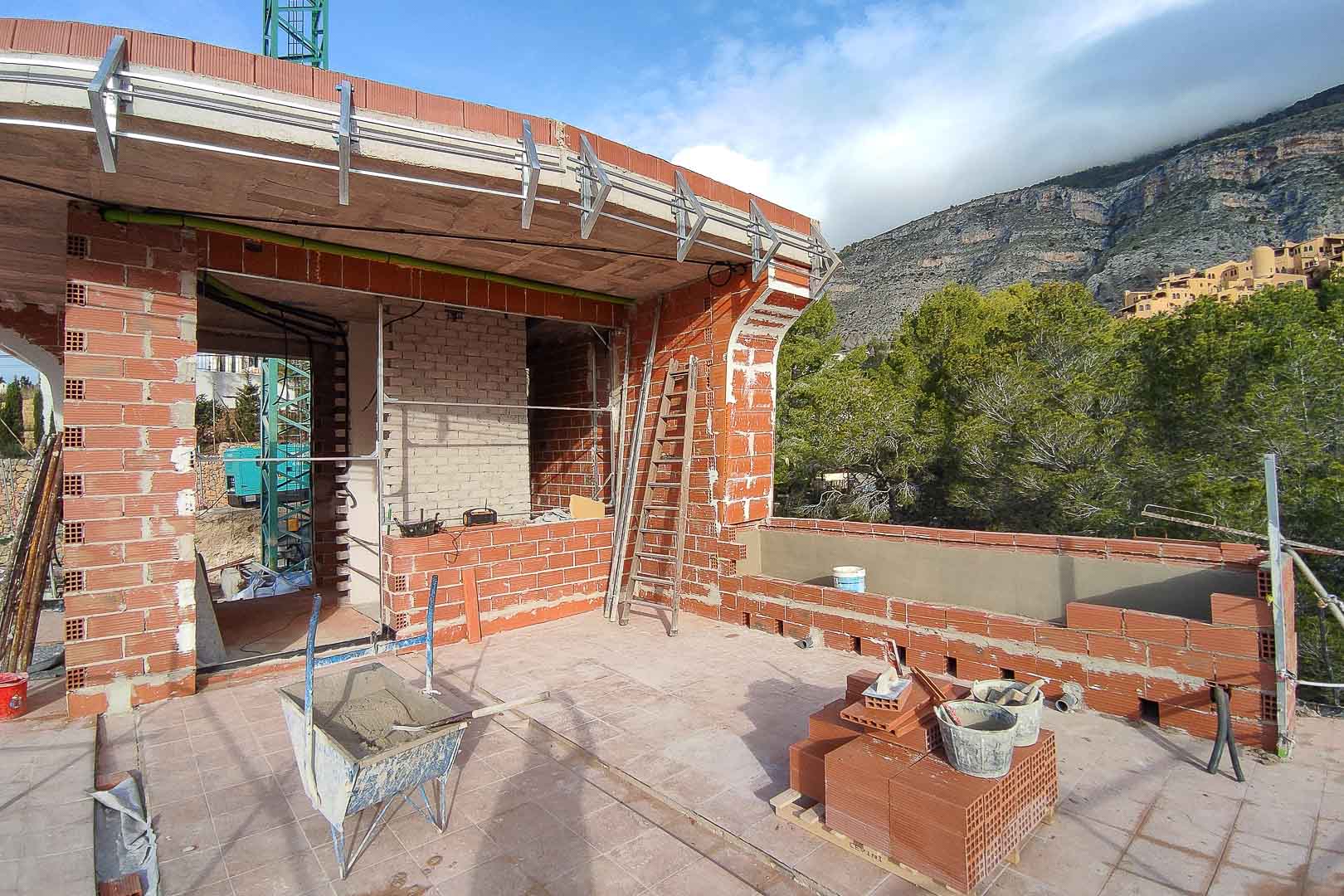 Phase de construction de la terrasse supérieure avec maçonnerie en briques rouges, dalles en béton et supports métalliques extérieurs offrant une vue sur la montagne.