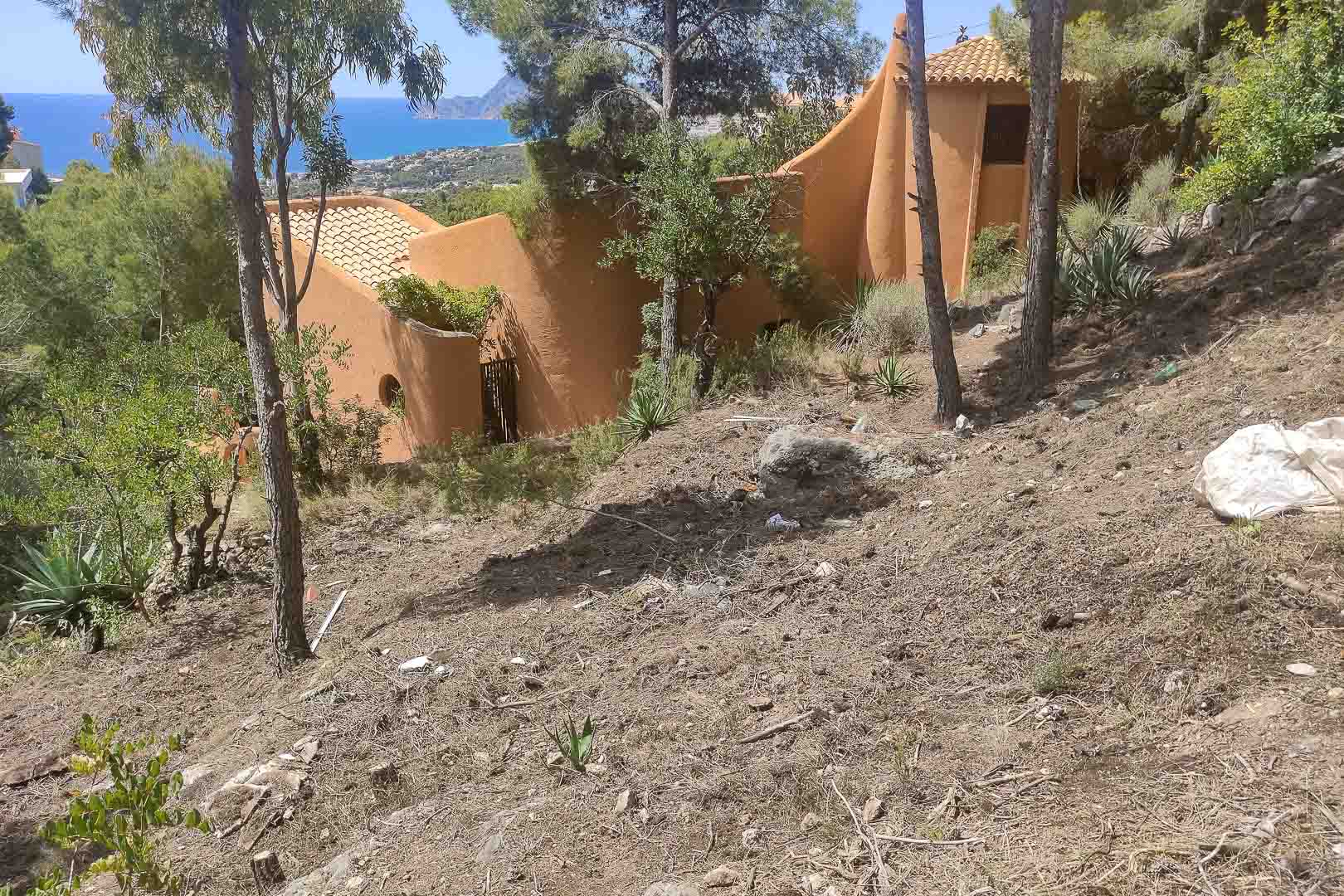 Vue extérieure de la villa à l'architecture organique et façades ocre, située sur un terrain en pente avec des pins et une vue panoramique sur la mer Méditerranée.