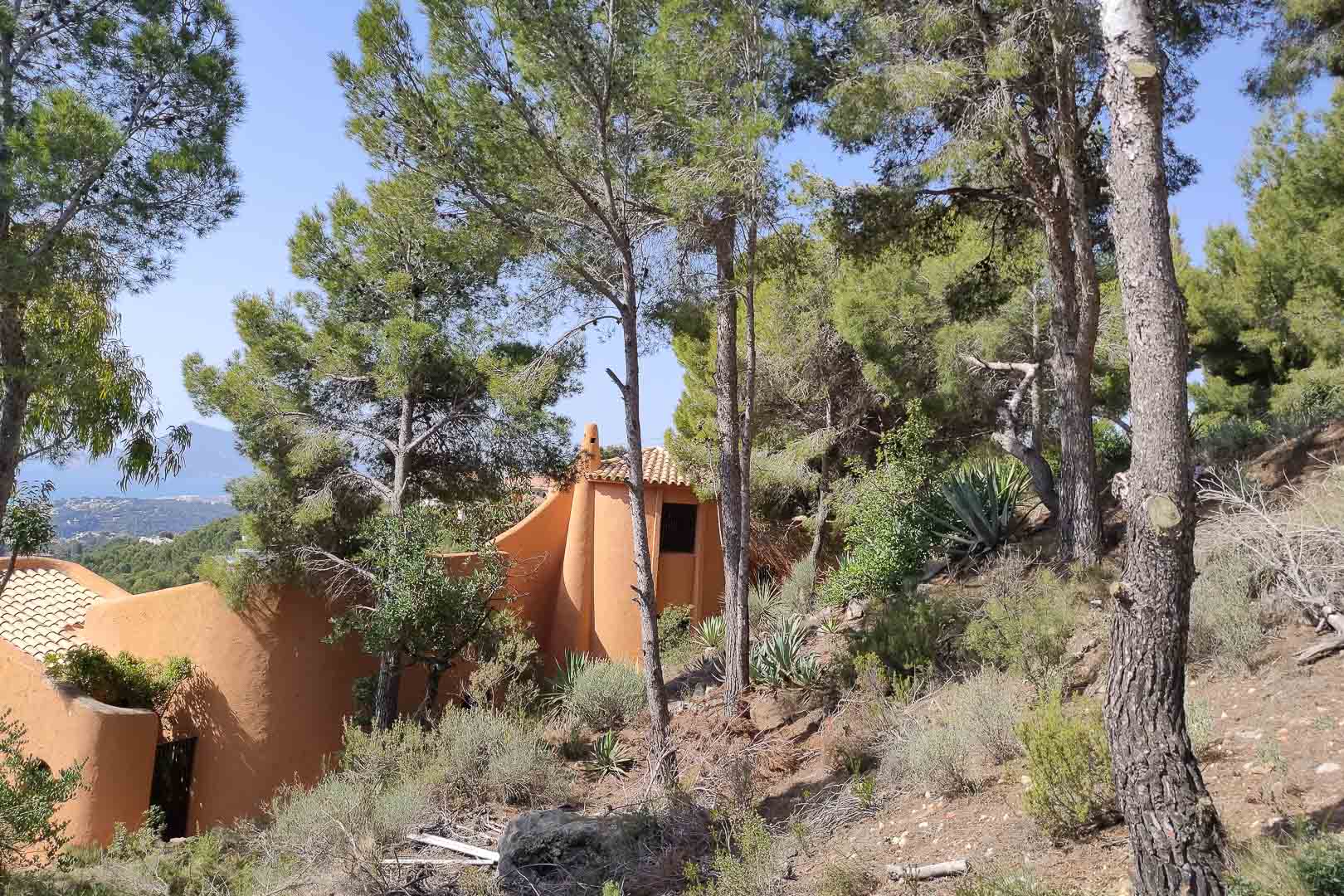 Vue extérieure de l'architecture organique aux tons terre cuite, entourée d'une pinède dense et d'une végétation méditerranéenne avec aperçu mer.