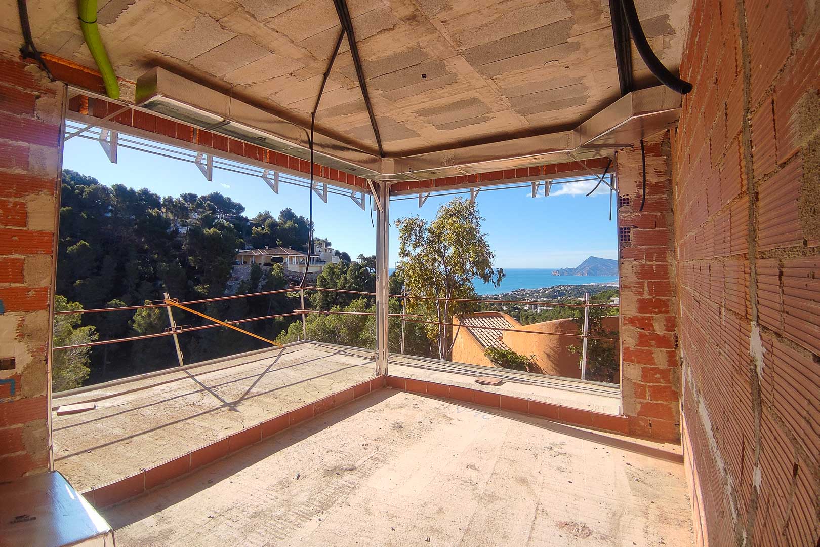 Vue panoramique sur la mer et la montagne depuis une pièce en construction avec de larges ouvertures d'angle et gaines techniques apparentes.