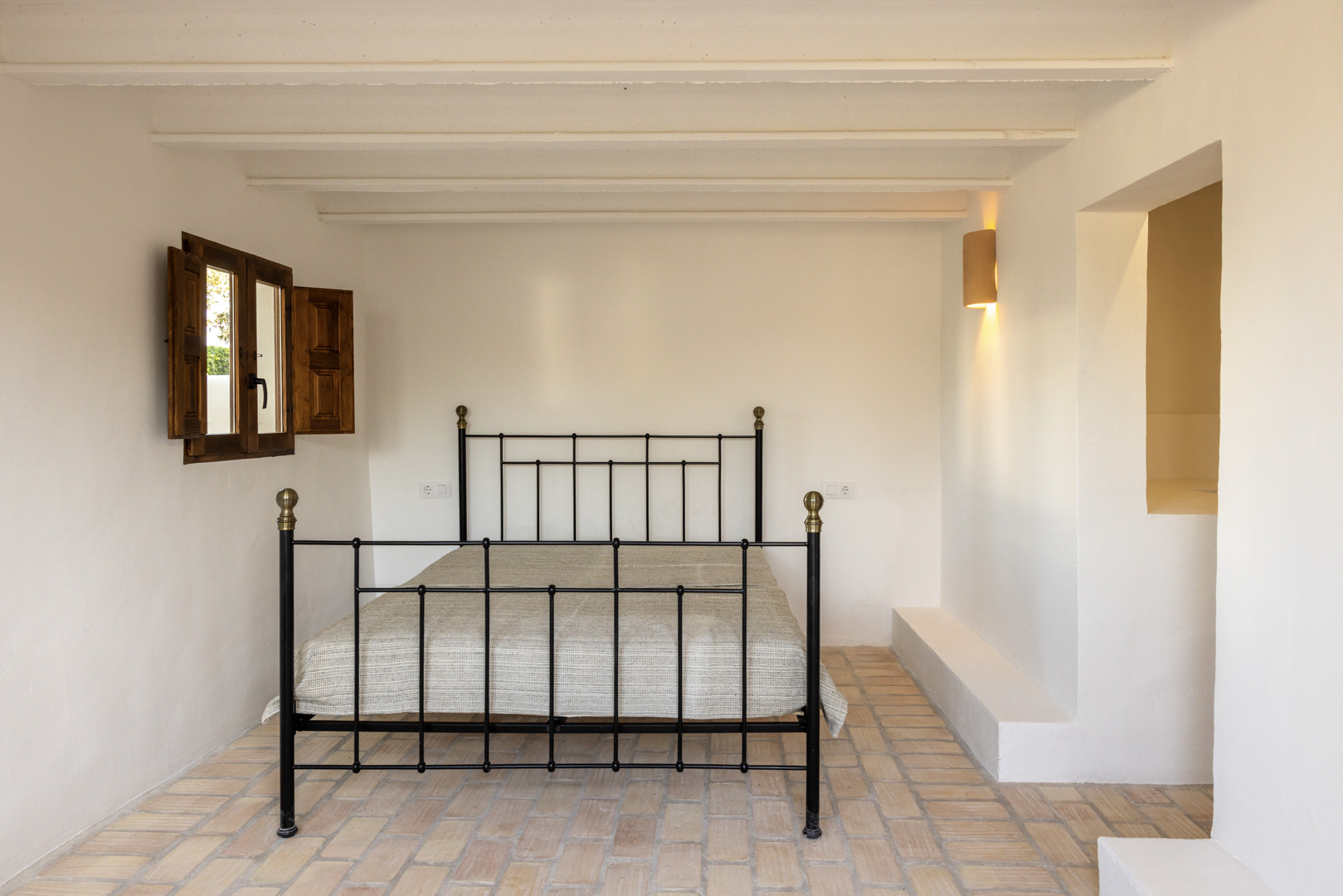Chambre lumineuse avec lit en fer forgé, volets en bois et sol en carreaux rustiques.
