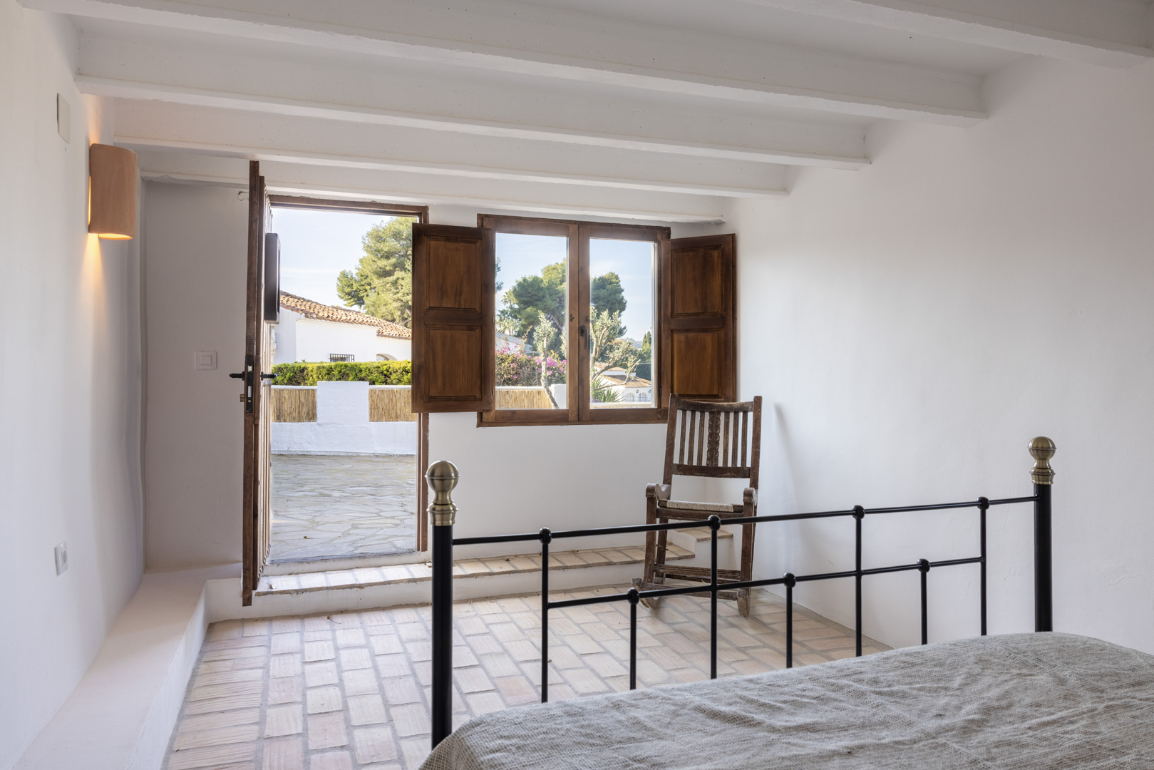 Chambre lumineuse avec sol en terre cuite, porte patio et fenêtre à volets en bois.