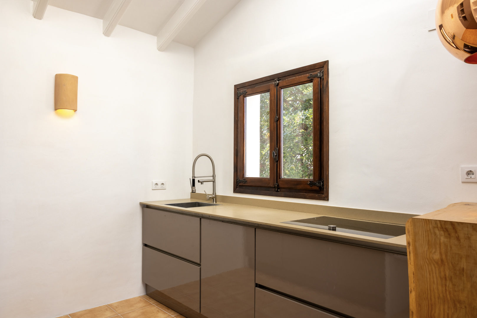 Détail de la cuisine avec comptoir beige, évier en acier et fenêtre en bois donnant sur l'extérieur.
