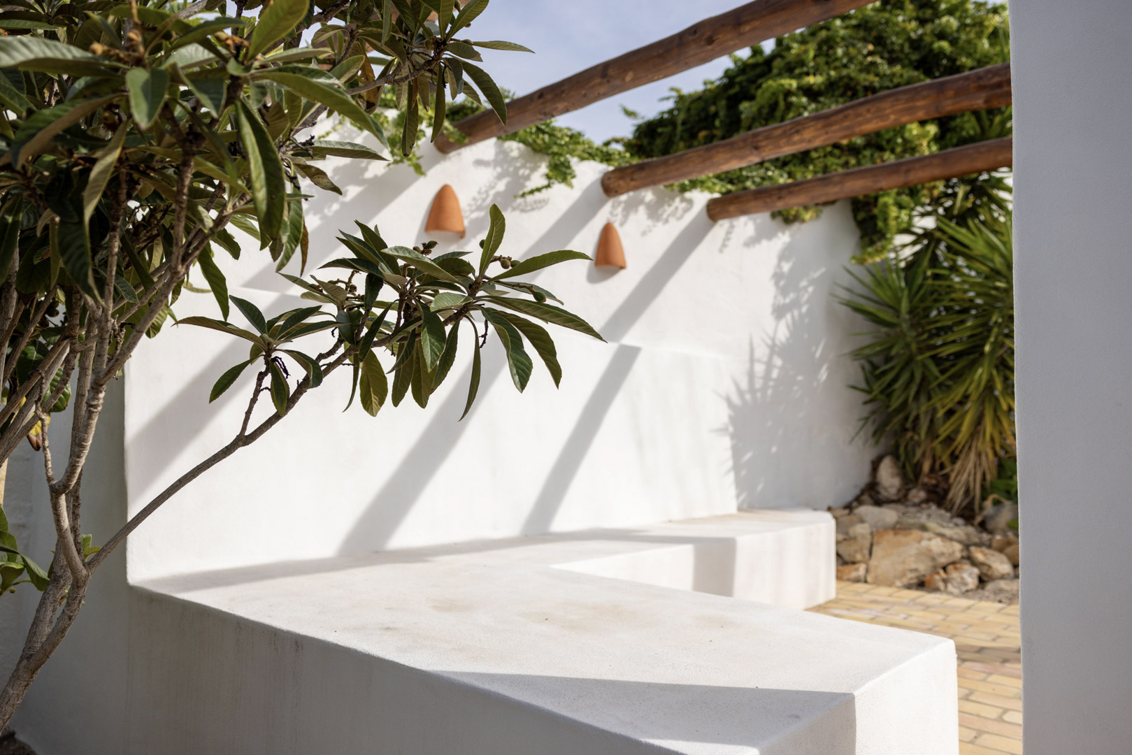 Détail de la terrasse avec banc en béton blanchi à la chaux, pergola en bois et végétation luxuriante.