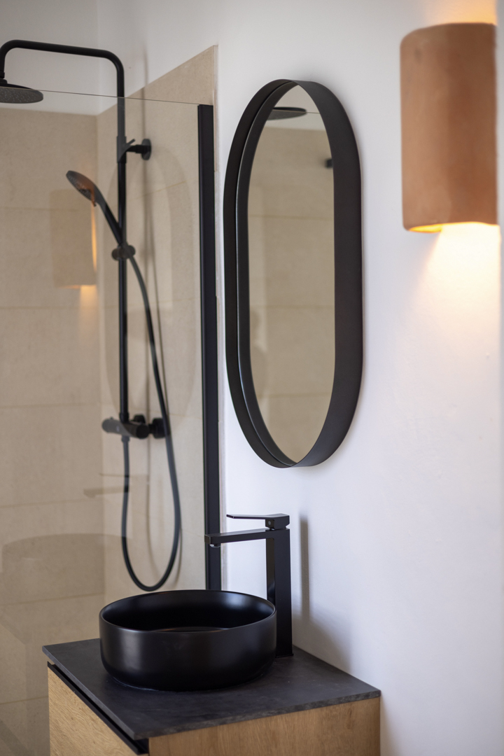 Détail de salle de bain avec lavabo noir circulaire, miroir ovale et douche vitrée.
