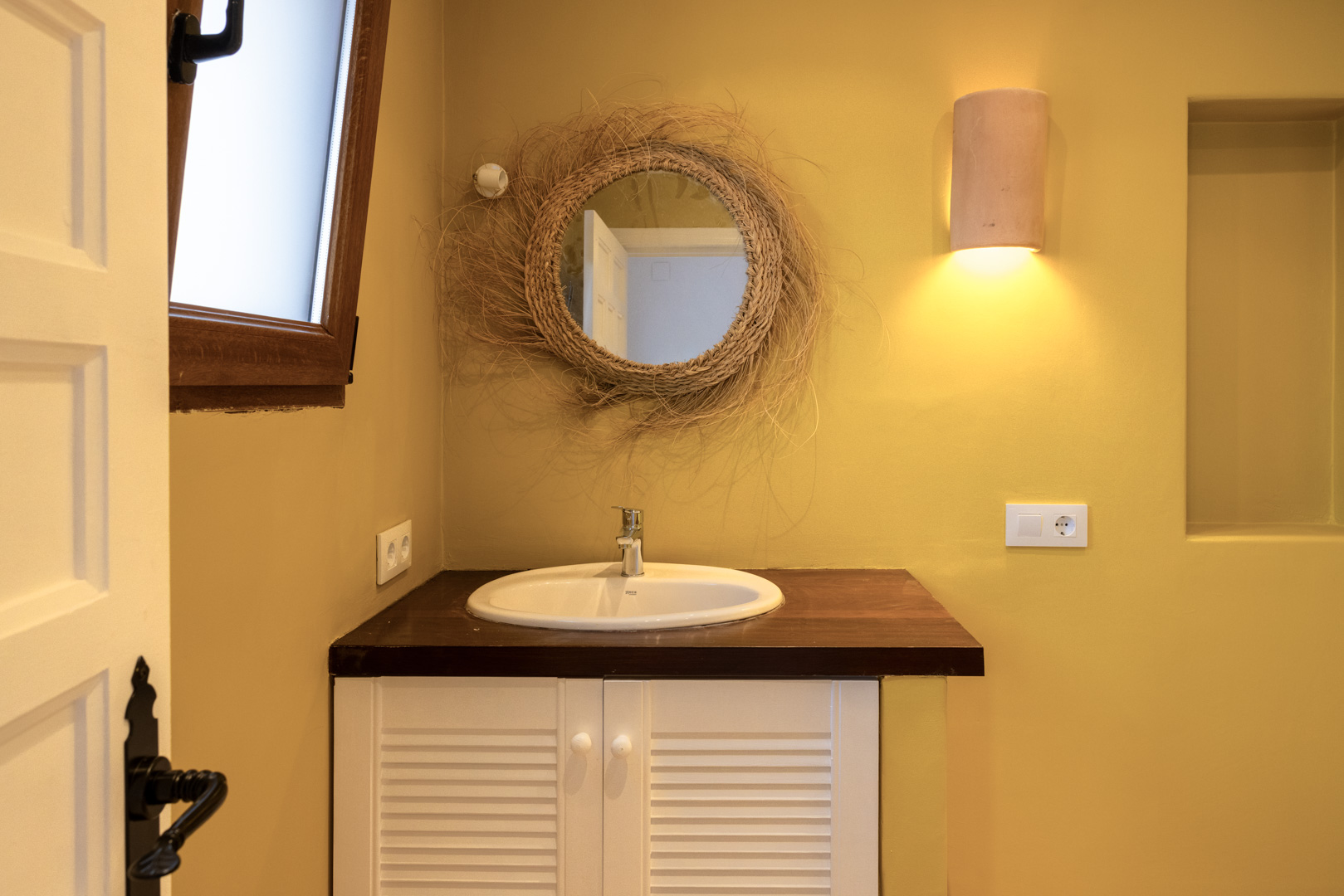 Détail de salle de bain avec lavabo ovale, miroir en osier et comptoir en bois foncé sur un meuble blanc.