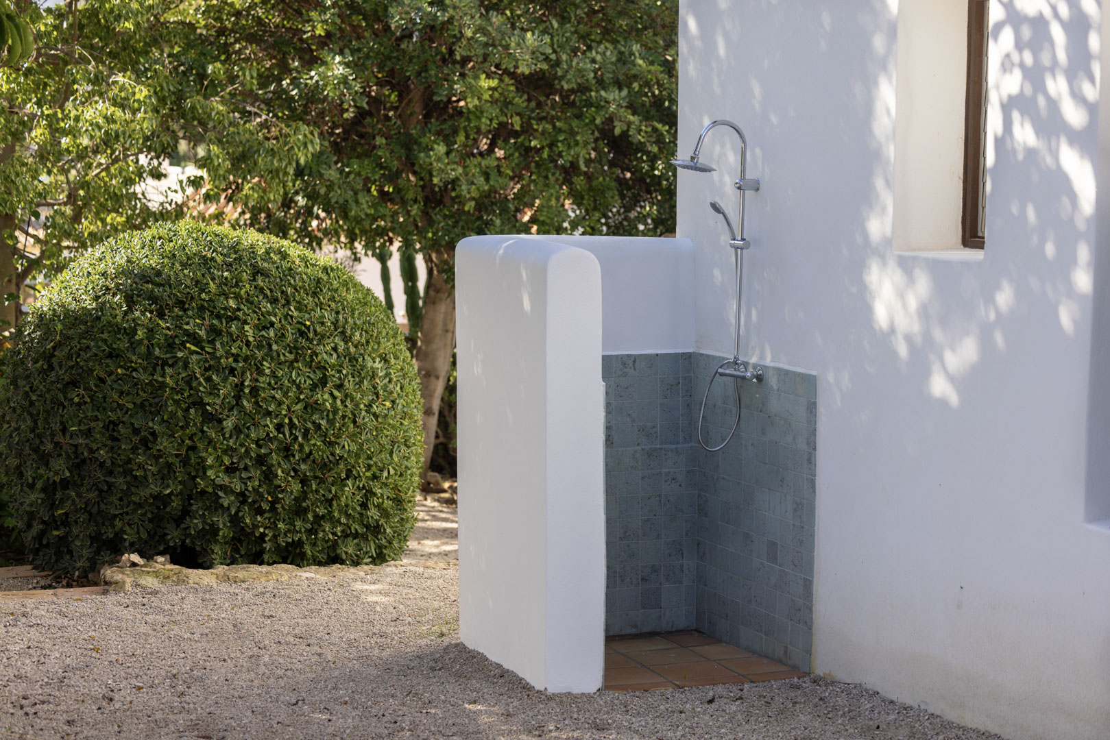 Détail d'une douche extérieure avec des carreaux gris et une base en terre cuite, à côté d'un jardin avec un arbuste sphérique.