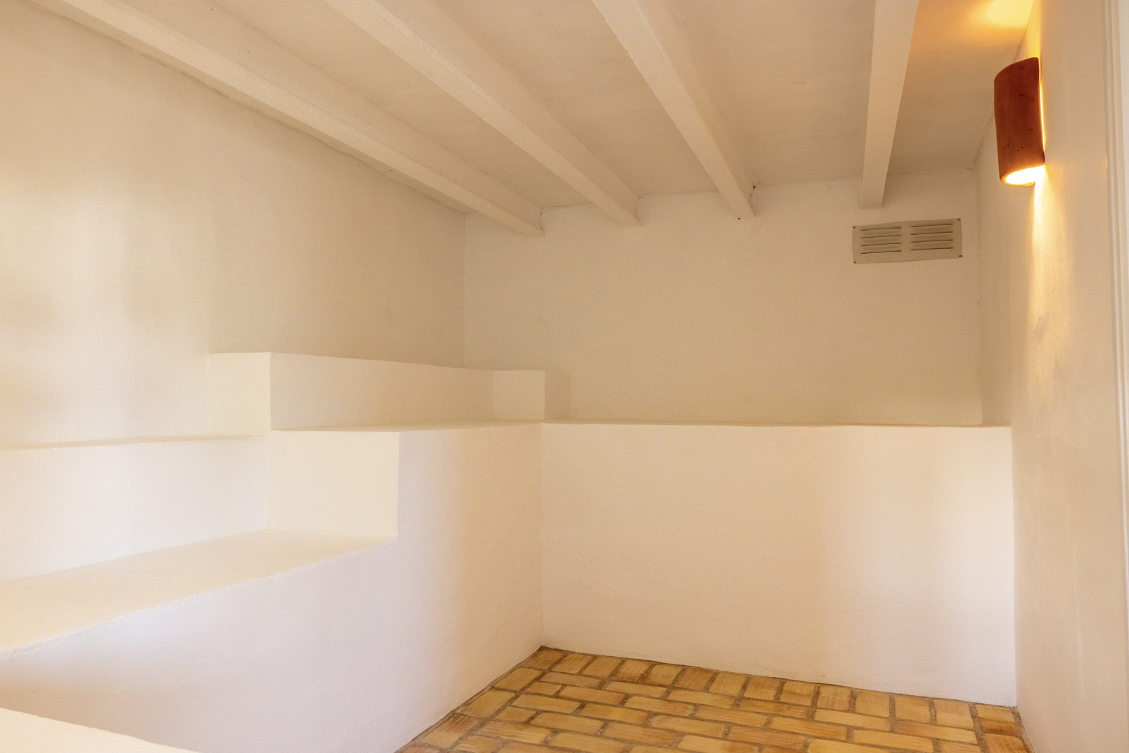Détail intérieur avec sol en brique, murs blancs et lumière chaude. Poutres apparentes au plafond.