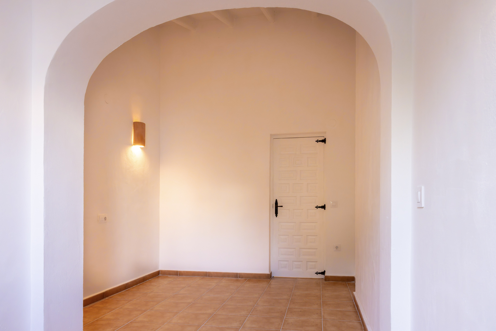 Entrée lumineuse avec arche, carrelage rustique et porte blanche à ferrure noire. Applique murale cylindrique.
