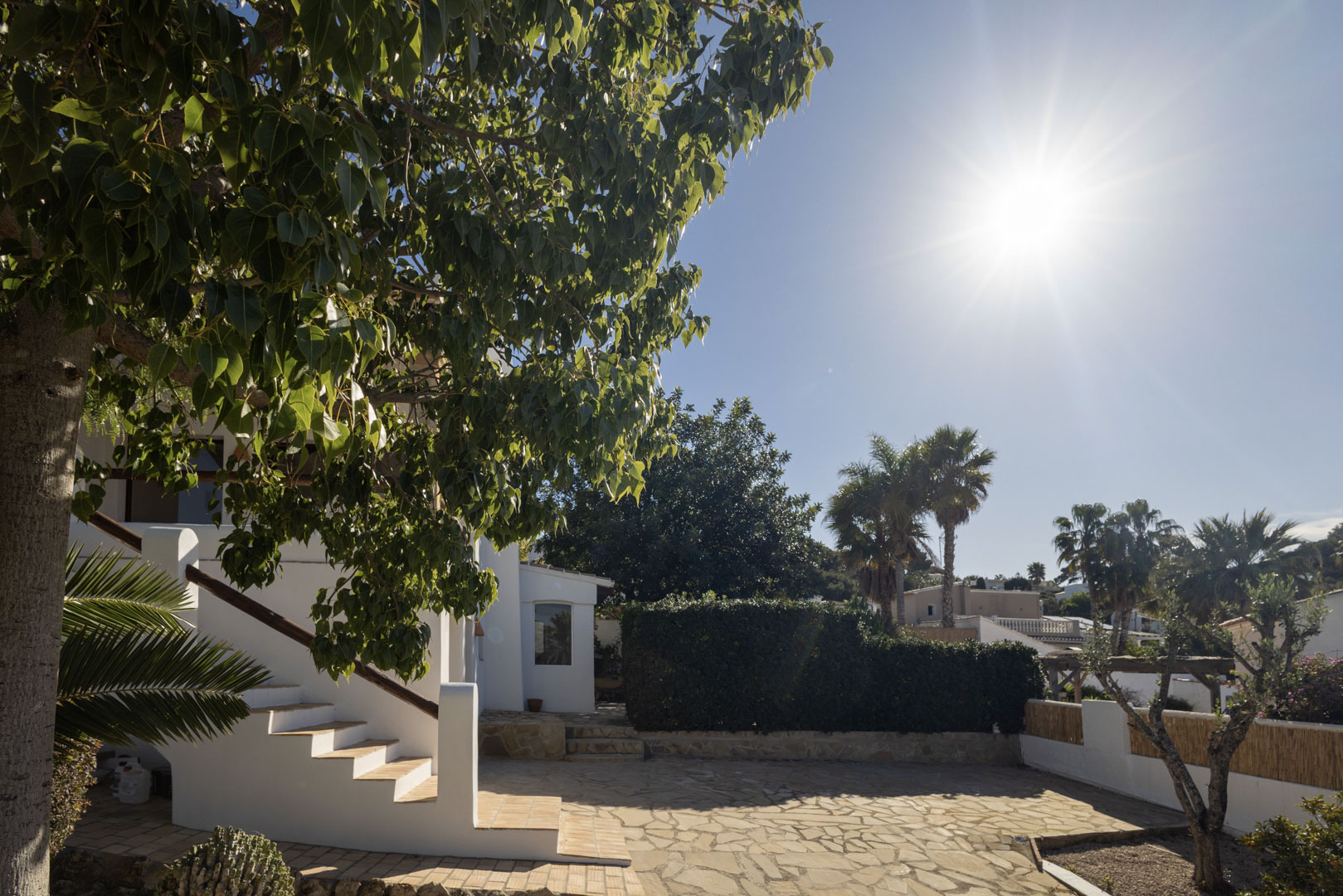 Extérieur de la villa avec patio en pierre, escaliers blancs et végétation luxuriante sous le soleil.