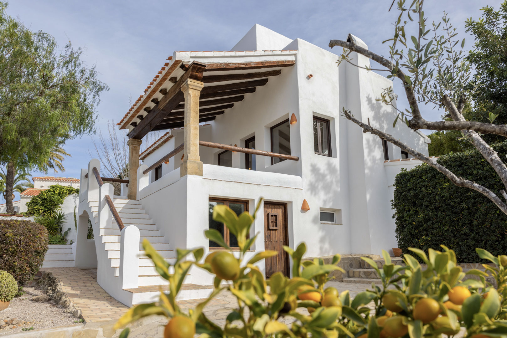 Vue extérieure de la villa à Jávea, mettant en valeur la façade blanche avec terrasse couverte aux poutres en bois et accès par escalier.