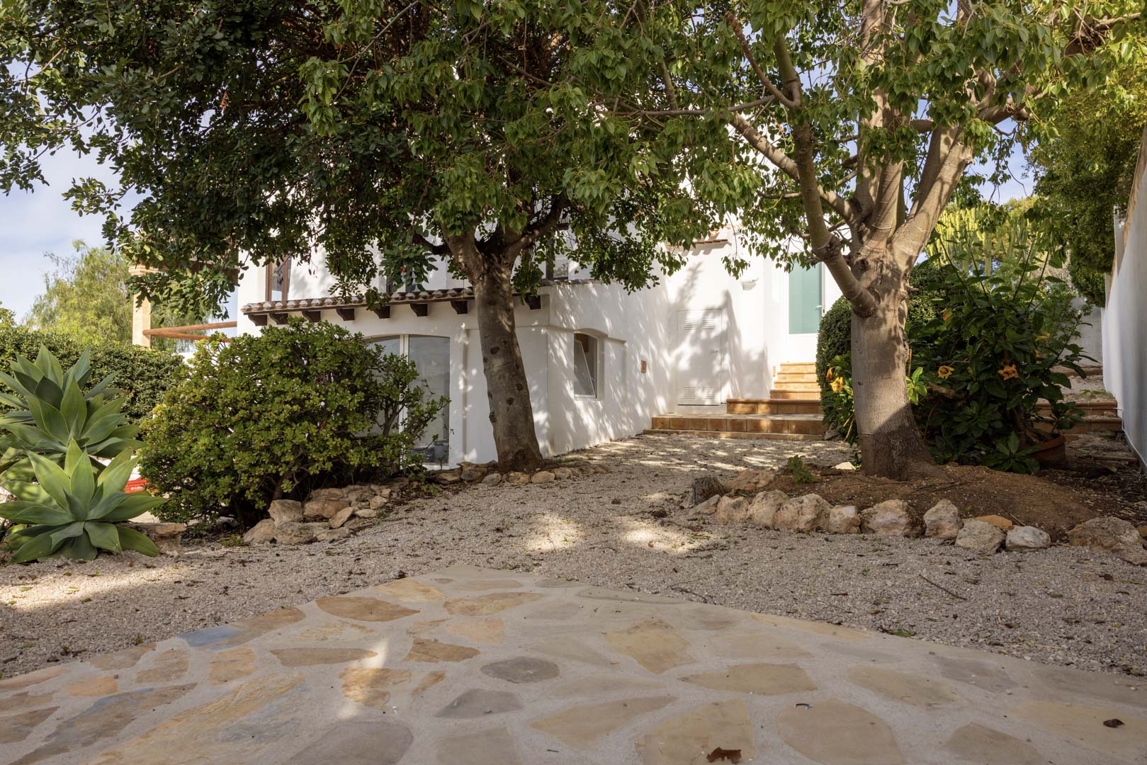 Vue extérieure de la villa avec jardin de gravier et accès à l'entrée. Végétation luxuriante et façade blanche.