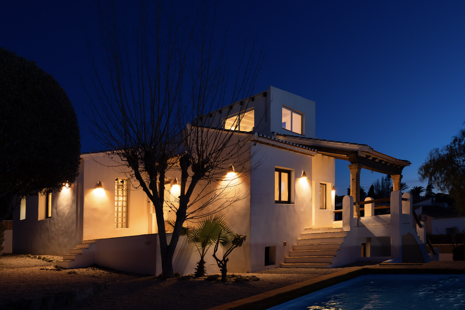 Vue nocturne de la villa illuminée avec piscine réfléchissante et façade blanche traditionnelle.