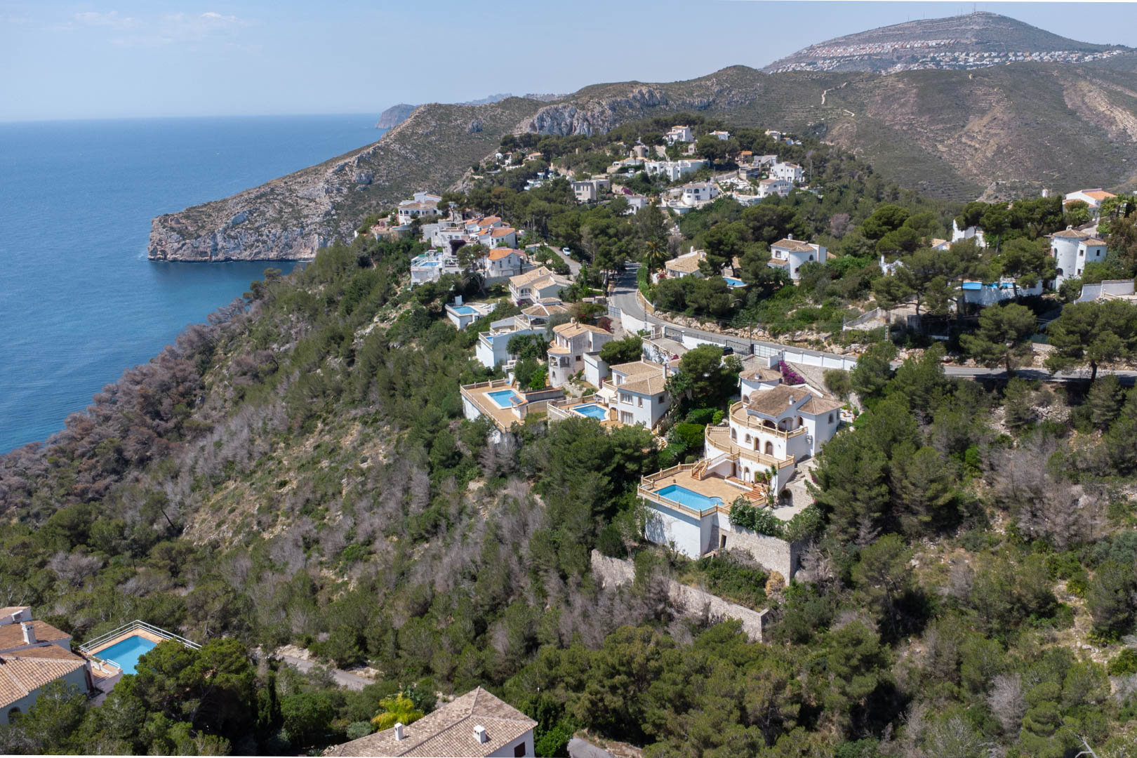 Vue aérienne de l'urbanisation Costa Nova à Jávea, présentant des villas blanches avec piscines privées situées sur une colline boisée face à la mer Méditerranée.