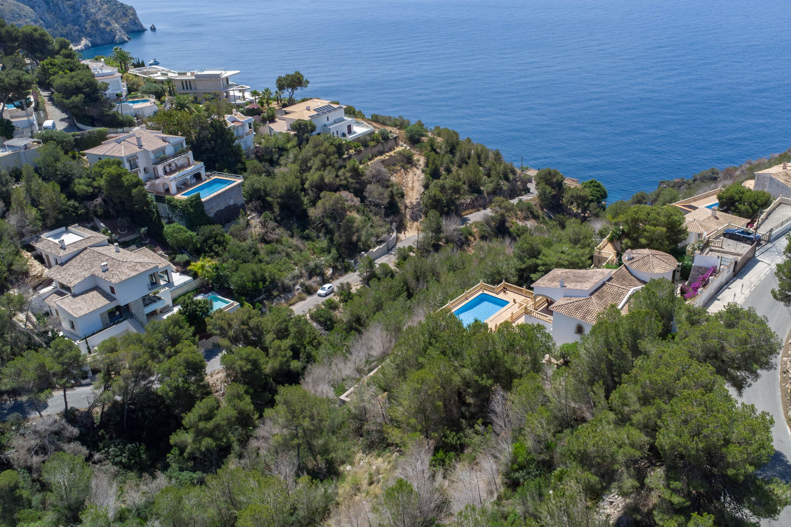 Vue aérienne des parcelles à Costa Nova, Jávea, entourées de pins méditerranéens avec des villas disposant de piscines et une vue dégagée sur la mer.