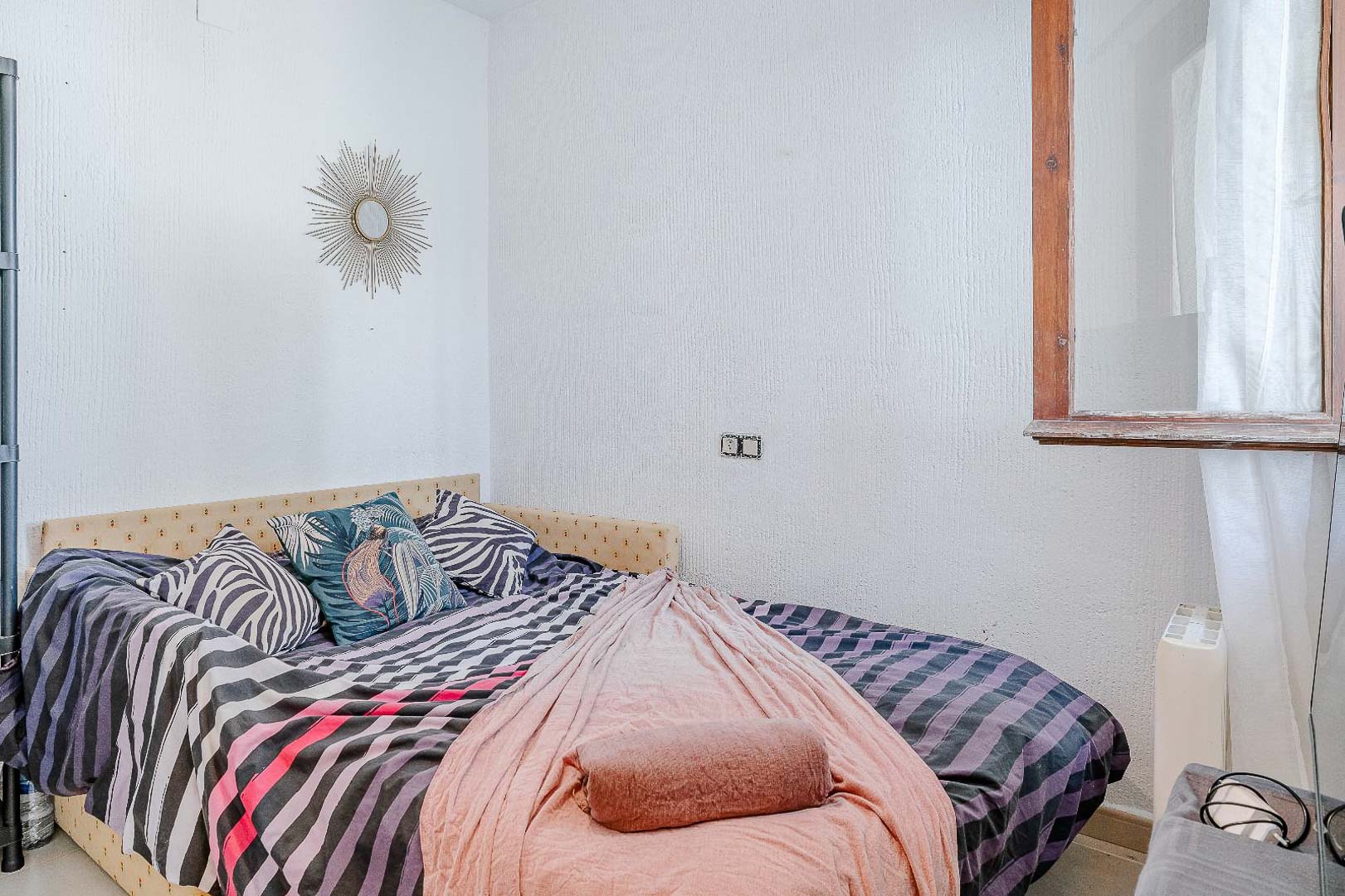 Chambre lumineuse avec murs texturés, lit simple avec tête de lit rembourrée et fenêtre à cadre en bois offrant une lumière naturelle constante.