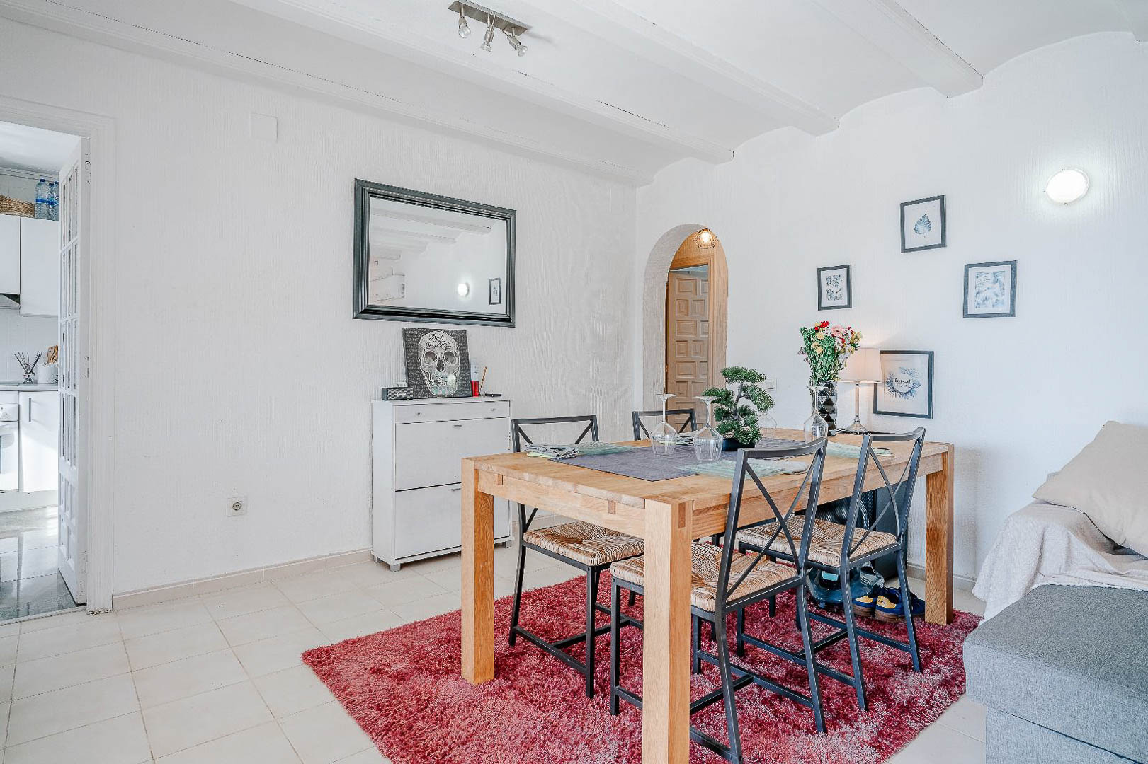 Coin repas avec table en bois massif et chaises en métal sur tapis rouge, caractérisé par des plafonds à poutres blanches et des ouvertures en arche.