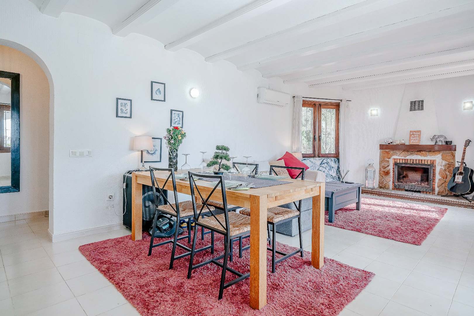Espace de vie ouvert comprenant un salon avec cheminée en pierre, des plafonds à poutres blanches et un coin repas avec table en bois et chaises en fer forgé.