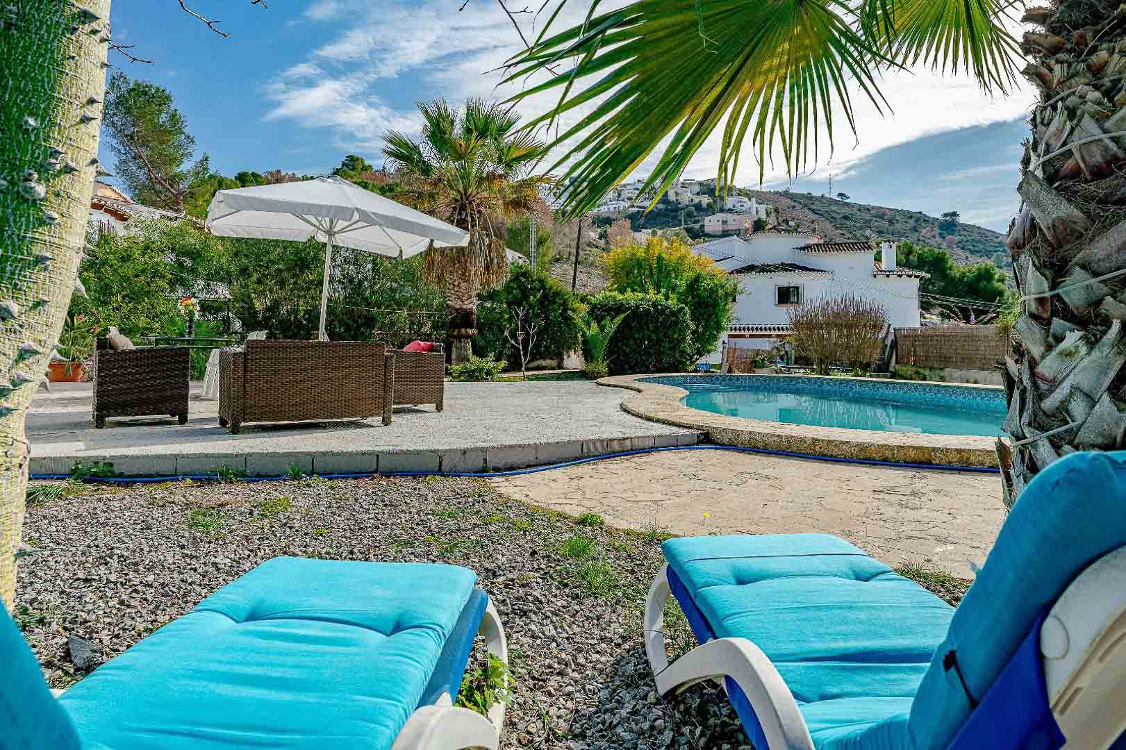 Espace piscine avec terrasse en pierre et gravier, comprenant des bains de soleil bleus et un salon de jardin en osier, entouré de palmiers et de végétation locale.