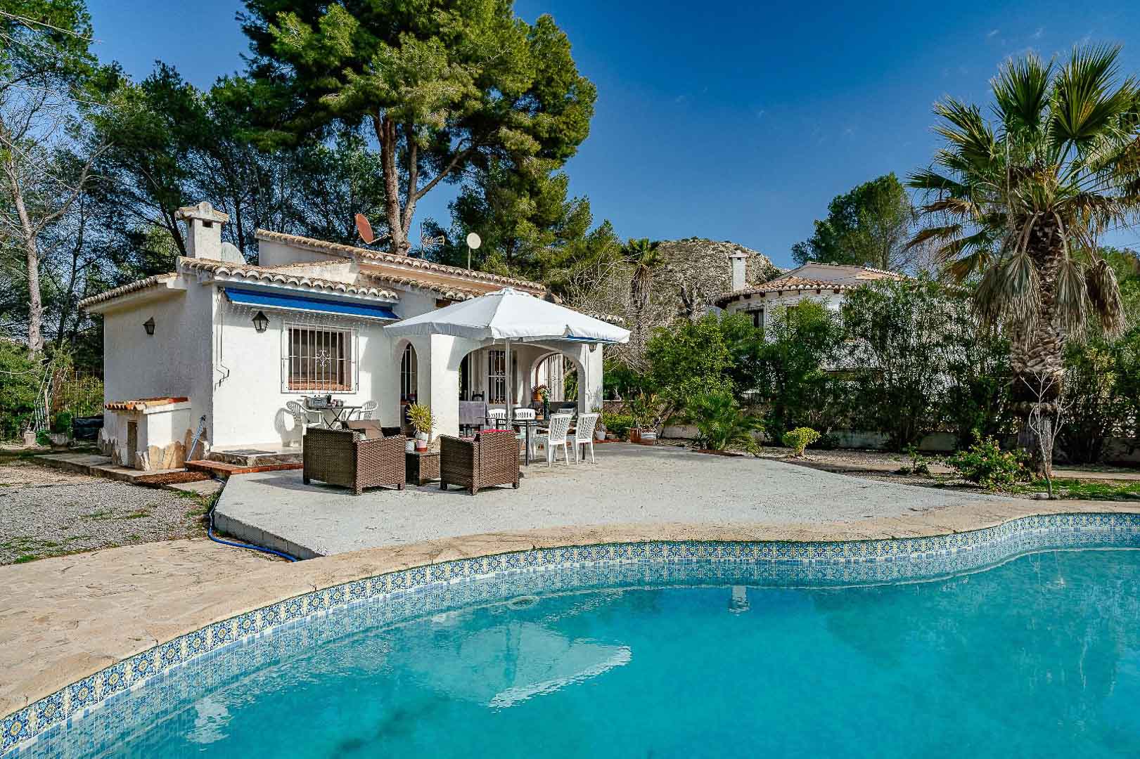 Extérieur de la villa avec piscine en mosaïque, vaste terrasse dallée et porche voûté traditionnel, entouré d'un jardin arboré de pins méditerranéens.