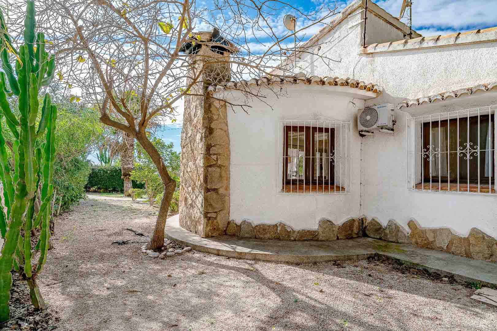 Façade méditerranéenne avec cheminée en pierre naturelle, fenêtres en bois protégées par des grilles blanches et jardin de gravier agrémenté de cactus et d'arbres.