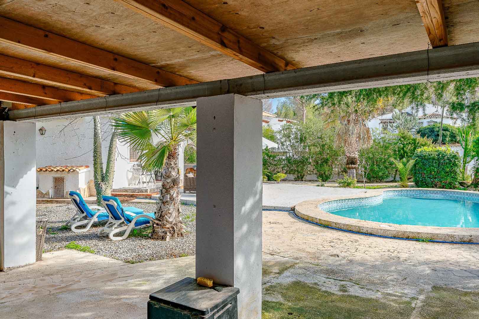 Porche couvert avec poutres en bois apparentes, offrant une vue sur la piscine de forme libre et le jardin méditerranéen agrémenté de palmiers.