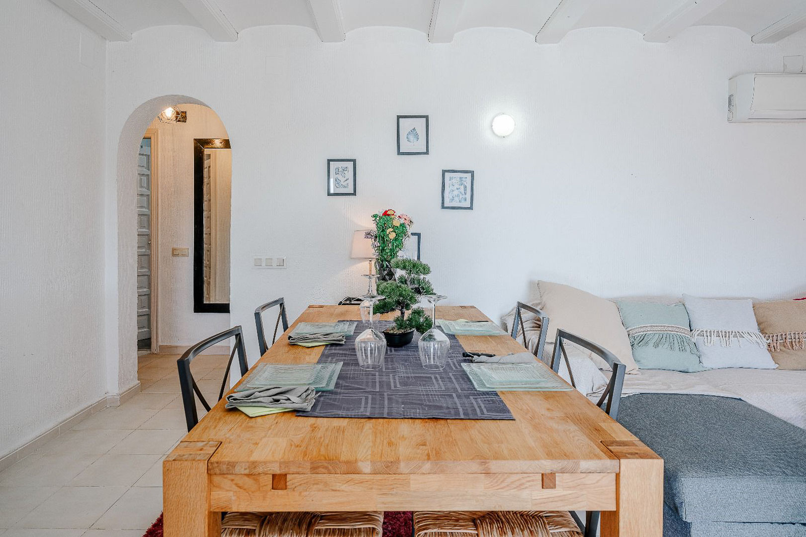 Salle à manger lumineuse avec table en bois massif et plafond à poutres blanches. Une arche mène au couloir à côté du coin salon avec canapé et climatisation.