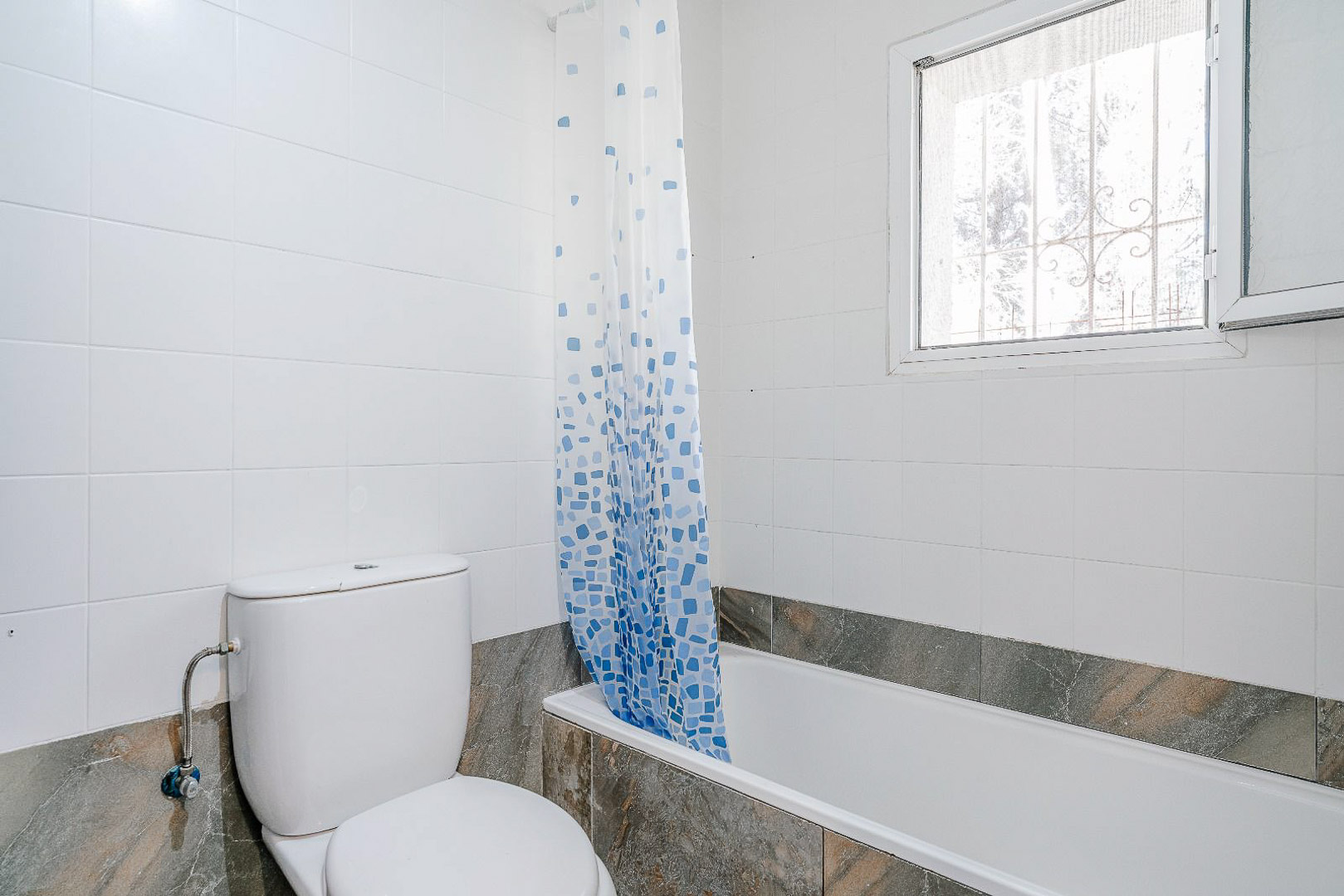 Salle de bain rénovée avec baignoire, toilettes en céramique blanche et carrelage mural clair. Une fenêtre avec grille assure la ventilation et la luminosité.