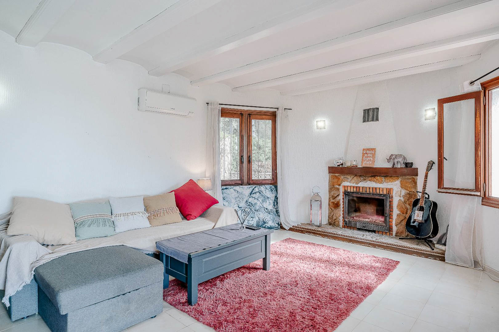 Salon lumineux avec plafond à poutres blanches, cheminée en pierre rustique et climatisation. Les fenêtres en bois offrent une belle lumière naturelle à cette pièce de vie.