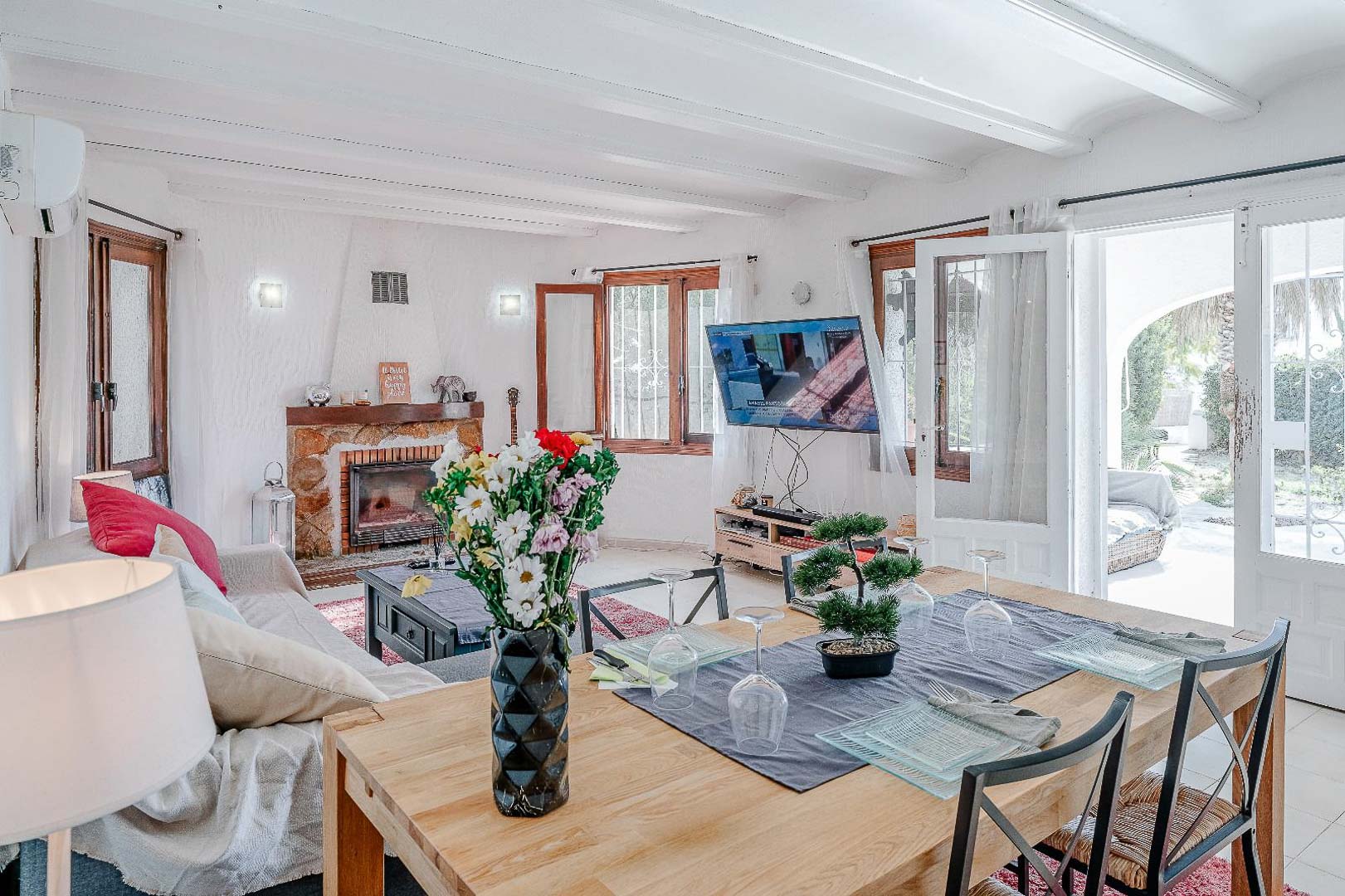 Salon-salle à manger avec plafonds à poutres blanches, cheminée en pierre naturelle et accès direct à la terrasse par des portes vitrées.