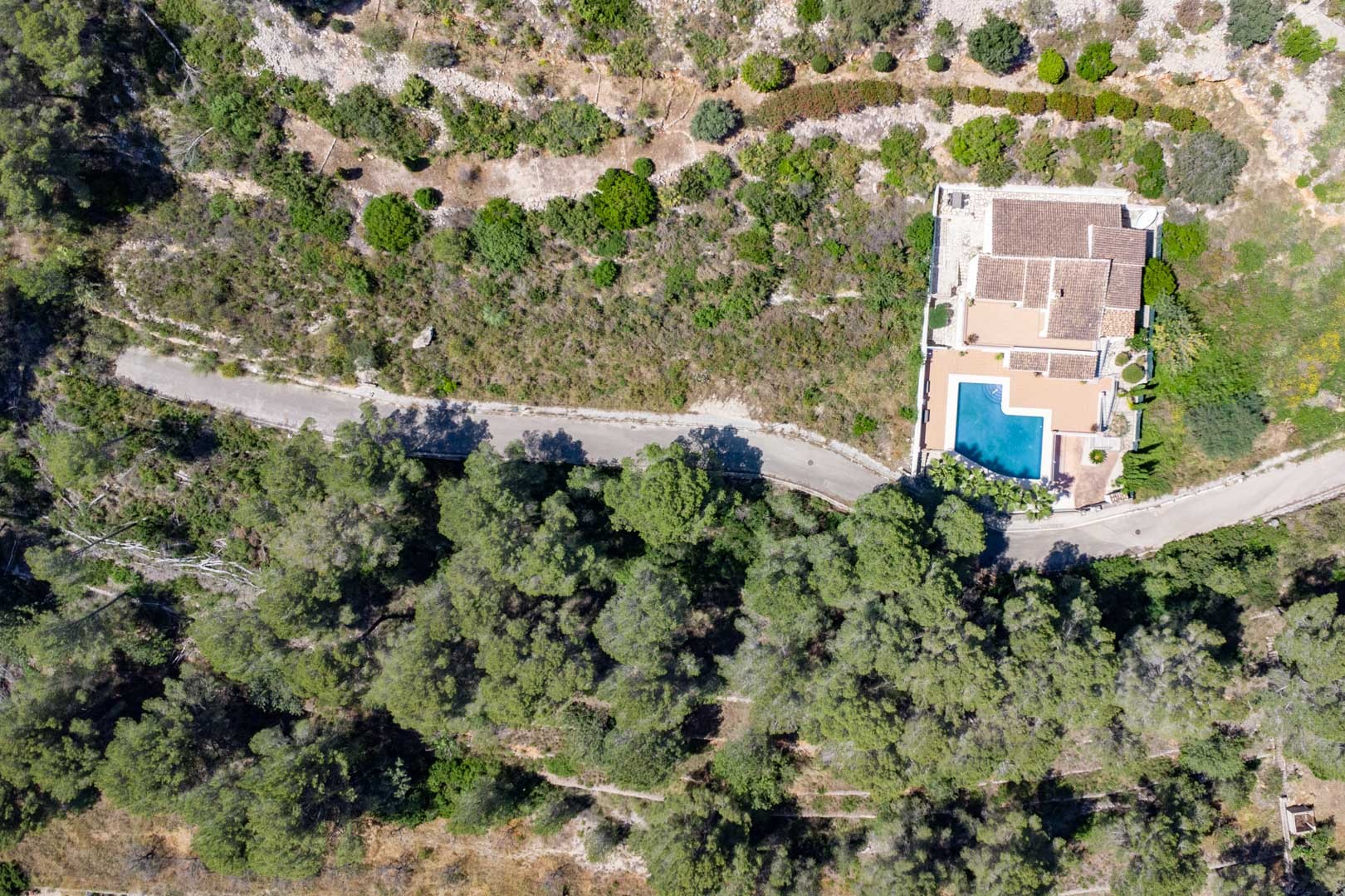 Vue aérienne d'une villa avec piscine en L et terrasse en terre cuite, entourée d'une forêt de pins dense sur les collines d'Alcalalí.