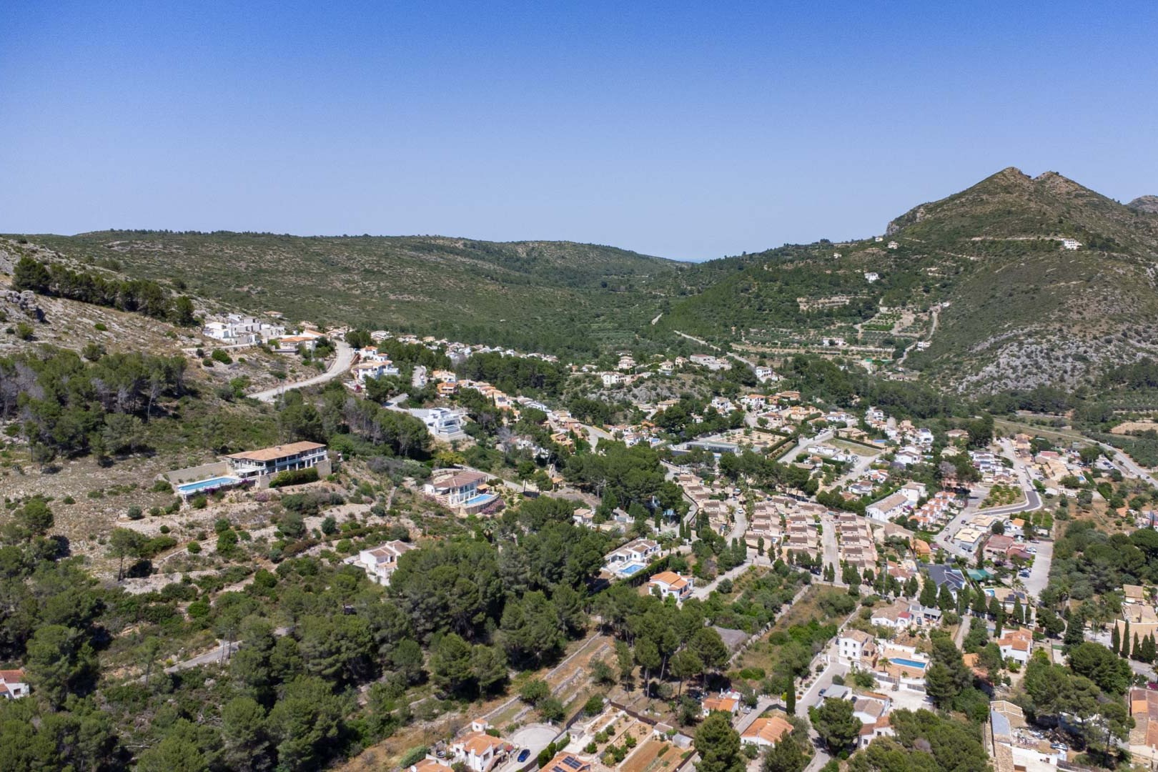 Vue aérienne panoramique d'Alcalalí, illustrant l'intégration des habitations sur le flanc de colline, entourées d'une végétation dense et de paysages montagneux.
