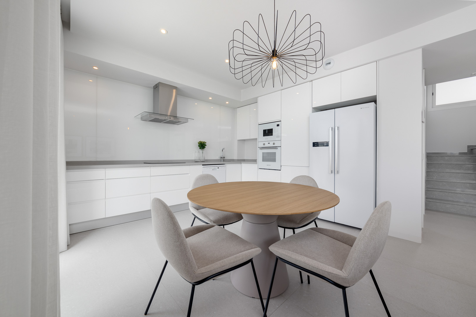 Cuisine minimaliste blanche avec électroménagers intégrés, plan de travail gris et coin repas avec table ronde en bois et luminaire design.