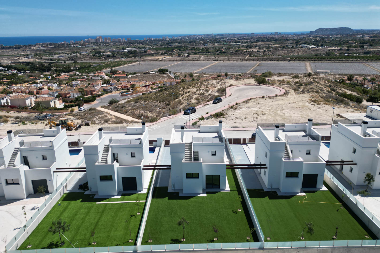 Vue aérienne de villas contemporaines aux façades blanches, avec jardins privés et solariums, offrant un panorama dégagé sur la côte et la mer au loin.