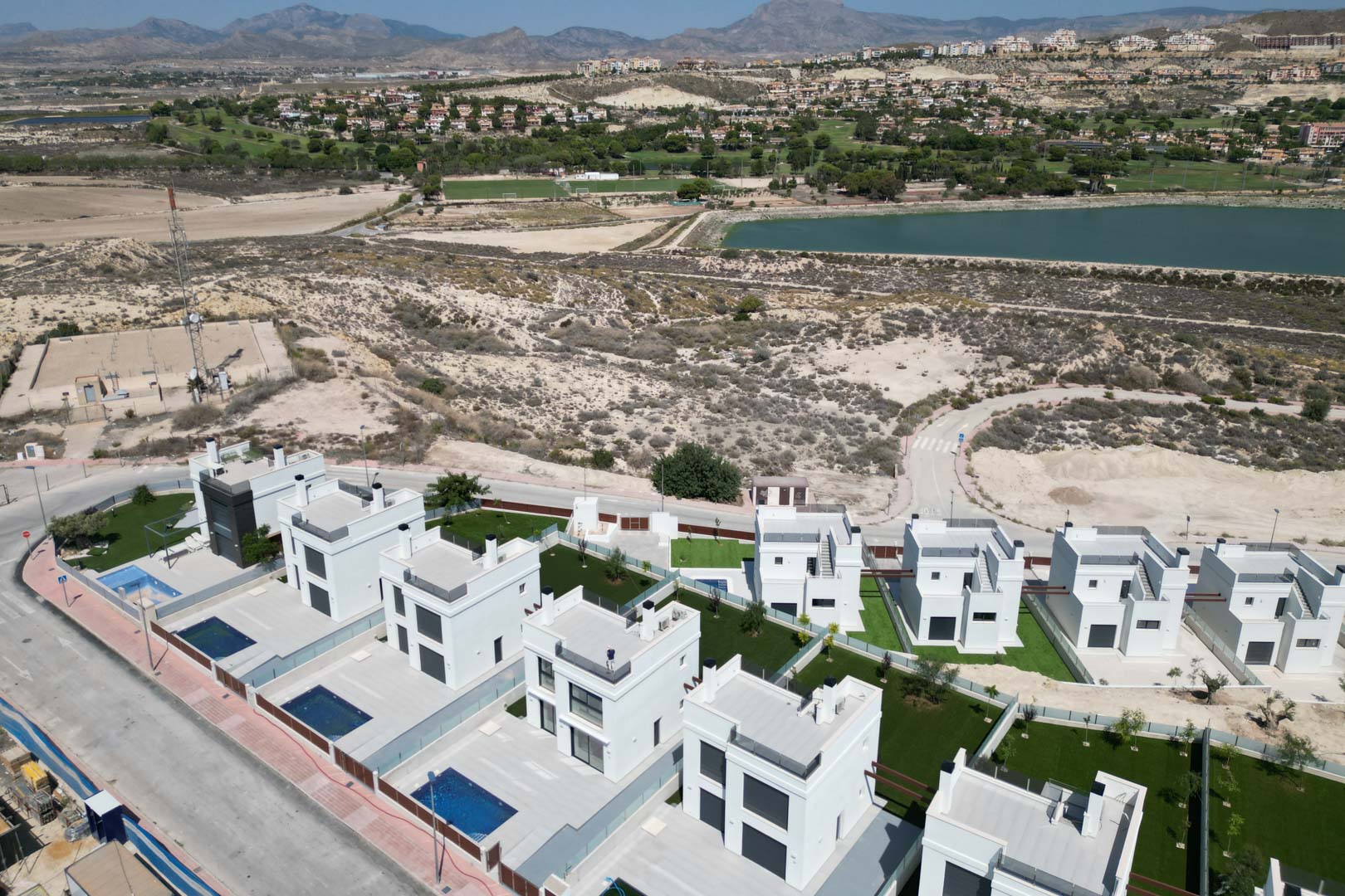 Vue aérienne de villas contemporaines blanches avec piscines privées et jardins, situées près d'un terrain de golf et d'un réservoir en montagne.