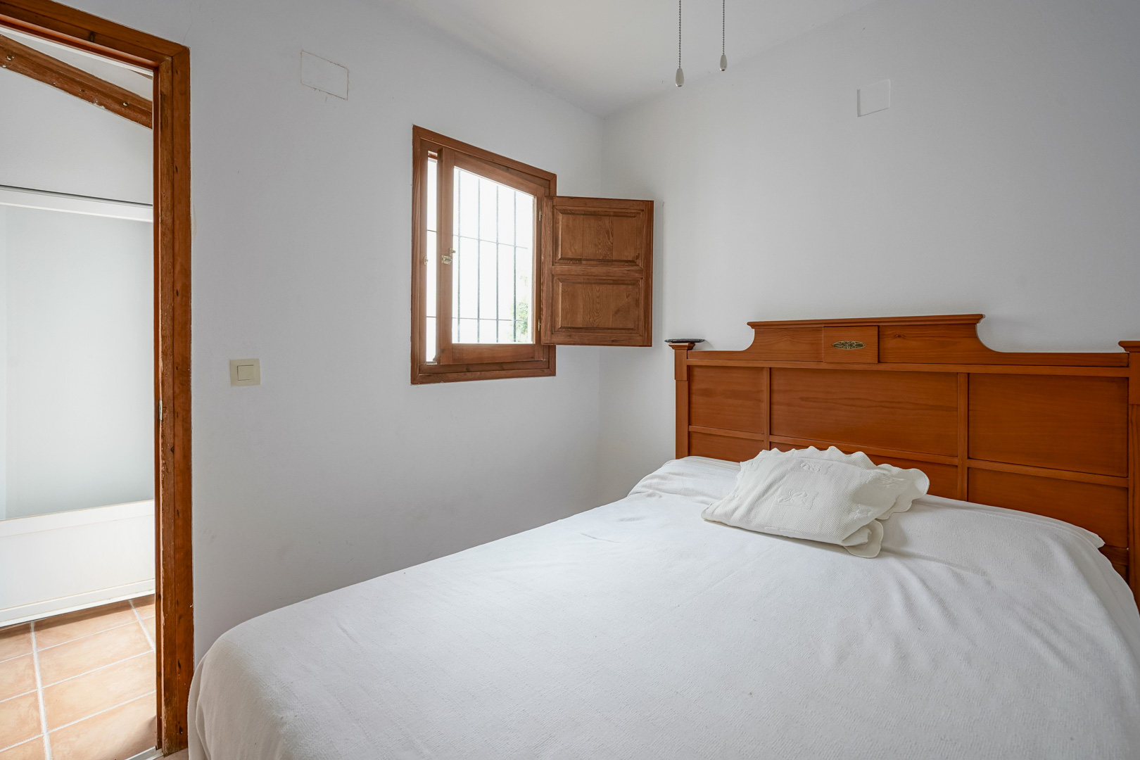 Chambre à coucher avec tête de lit en bois, fenêtre à volet intérieur traditionnel et vue sur une pièce attenante au sol en terre cuite.
