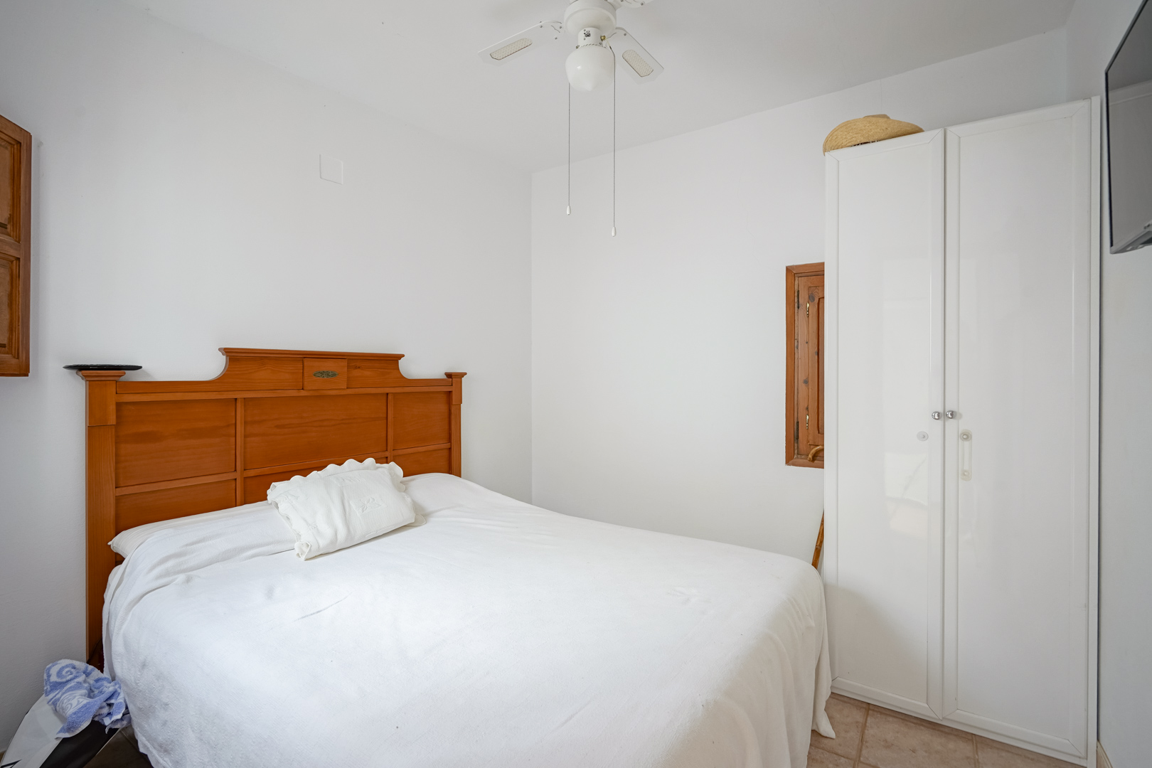 Chambre avec lit double, tête de lit en bois, armoire blanche et ventilateur de plafond. Les murs blancs et le sol en grès optimisent la clarté.