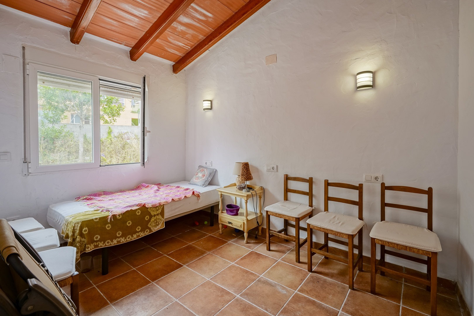 Chambre avec plafonds inclinés et poutres en bois apparentes, sol en terre cuite et murs blancs. Équipée d'appliques murales et d'une fenêtre donnant sur l'extérieur.
