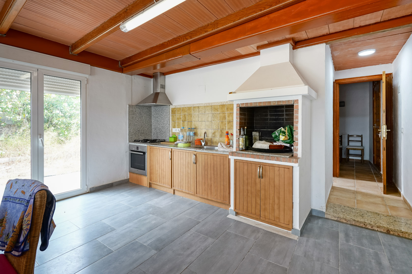 Cuisine fonctionnelle avec plafond en bois et poutres apparentes, comprenant un barbecue intégré, des plans de travail en granit et un accès extérieur.
