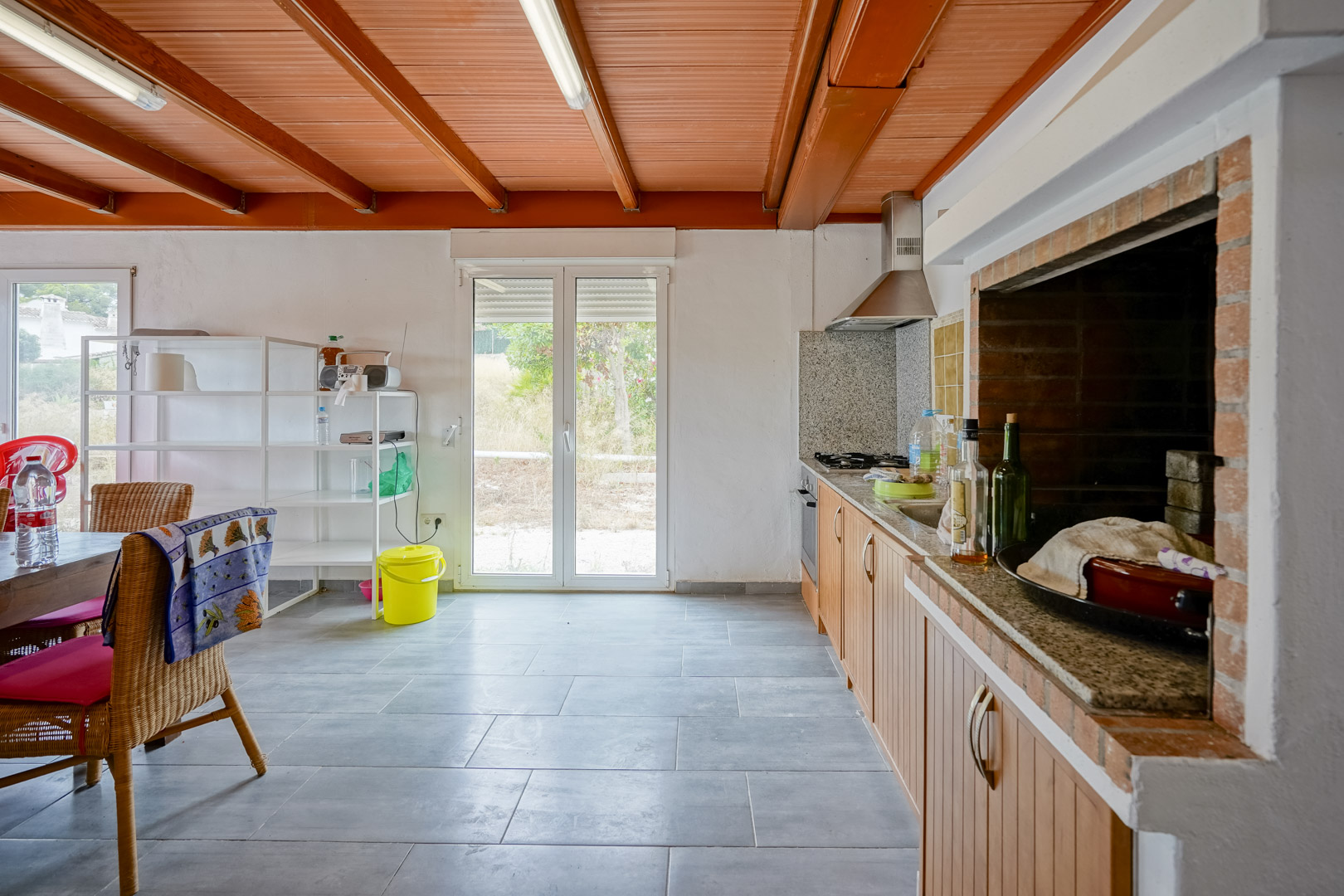 Cuisine rustique avec poutres apparentes en bois, plan de travail en granit et barbecue intégré en briques, offrant un accès direct au jardin par des portes vitrées.