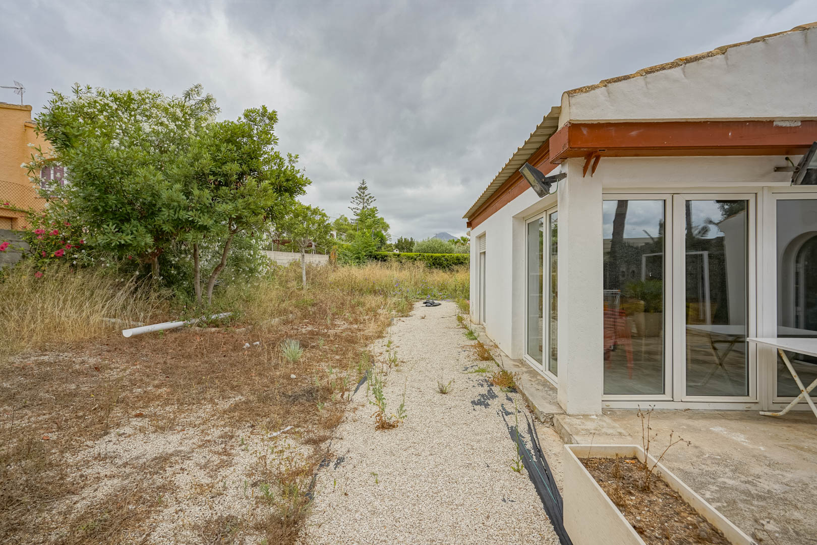 Façade latérale de la villa avec de grandes baies vitrées en PVC blanc ouvrant sur un vaste terrain naturel agrémenté de végétation méditerranéenne.