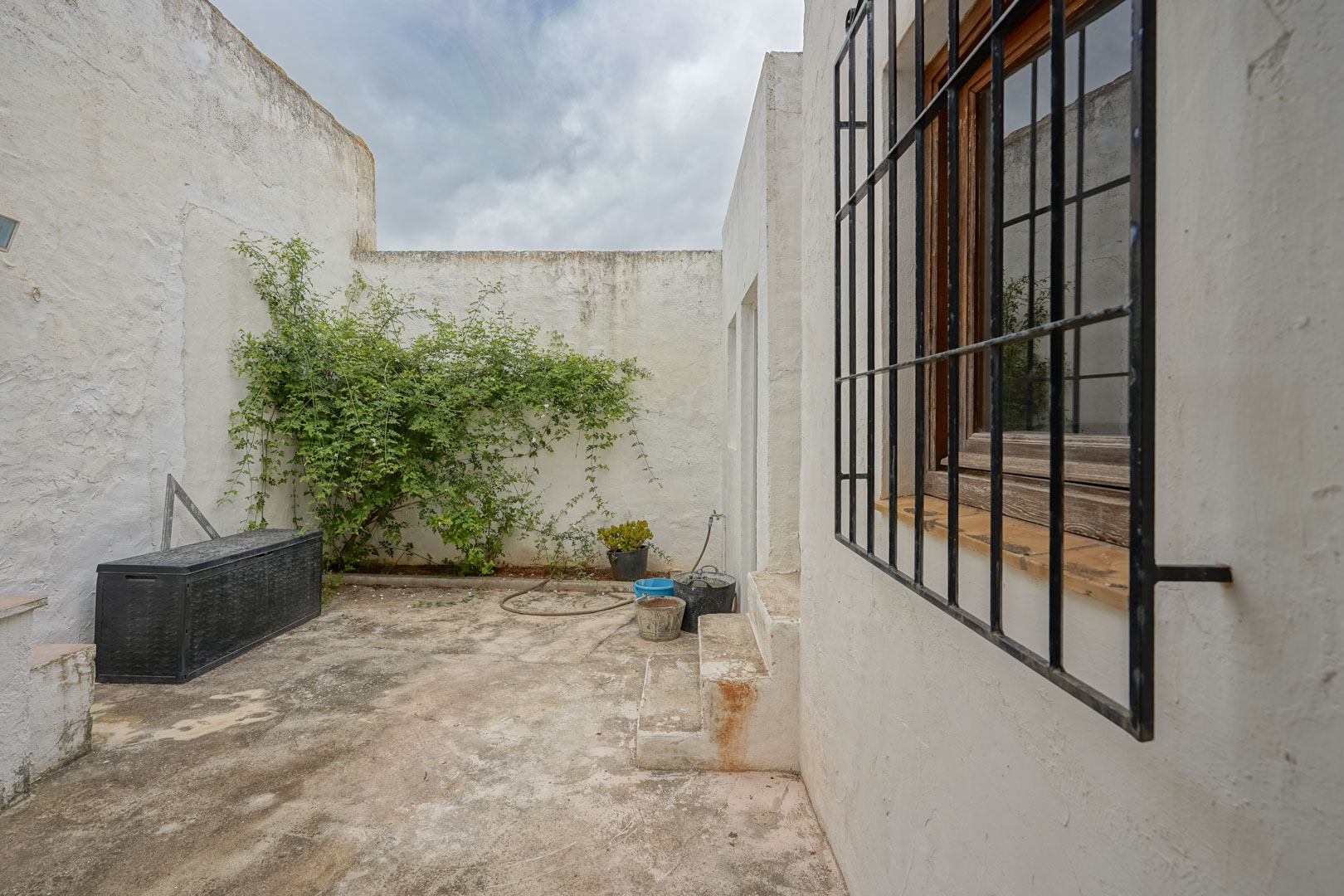 Patio intérieur avec murs blanchis à la chaux, sol en béton, fenêtre avec grille de sécurité en fer et espace de rangement extérieur.