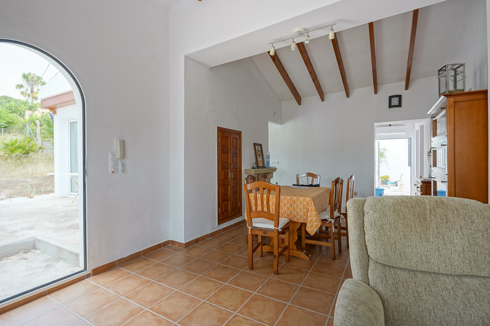 Salle à manger avec hauts plafonds à poutres apparentes, sol en terre cuite et porte vitrée en arche offrant un accès direct à l'extérieur.