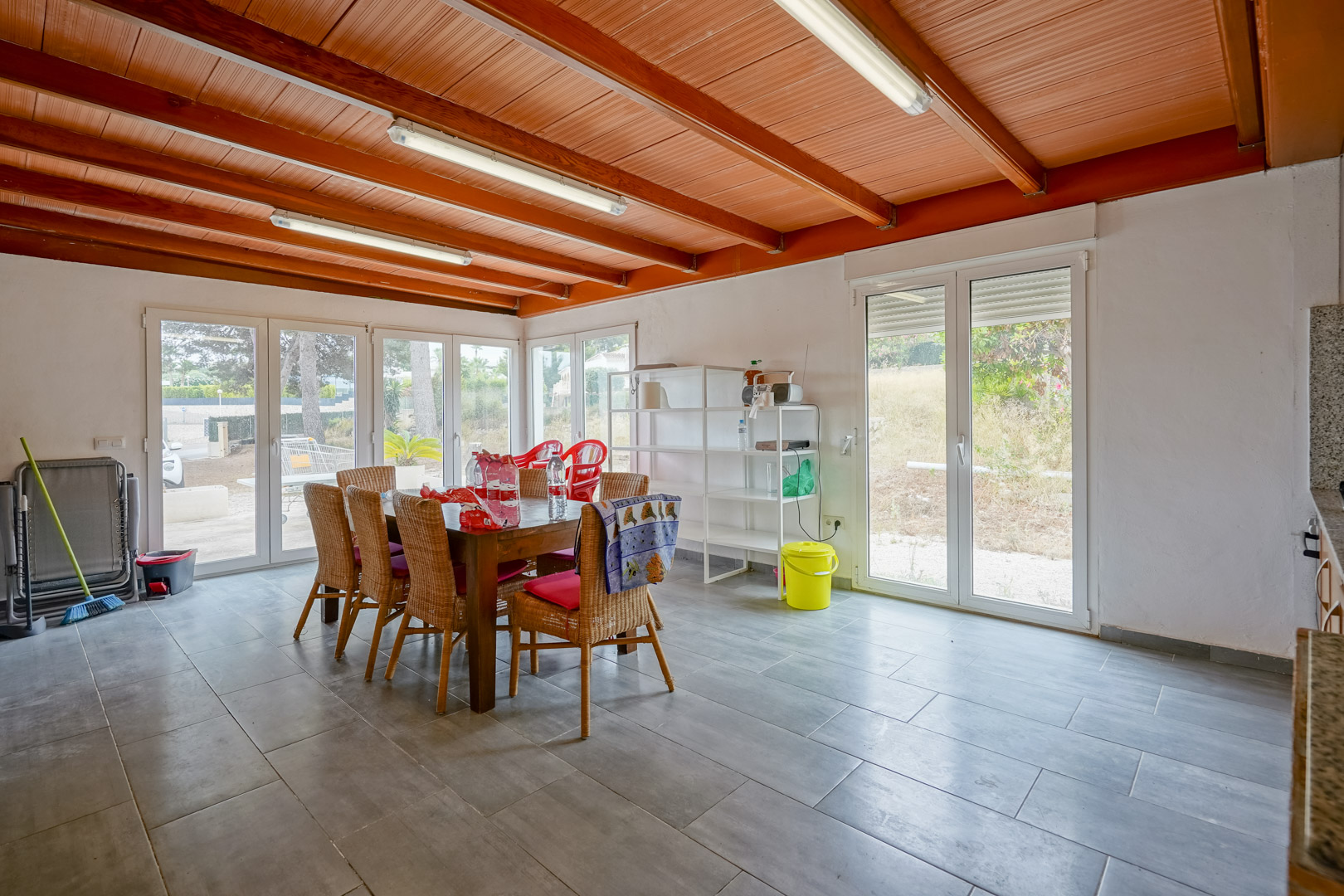 Salle à manger lumineuse avec plafond à poutres apparentes en bois, sol en grès gris et grandes baies vitrées offrant un accès direct à l'extérieur.