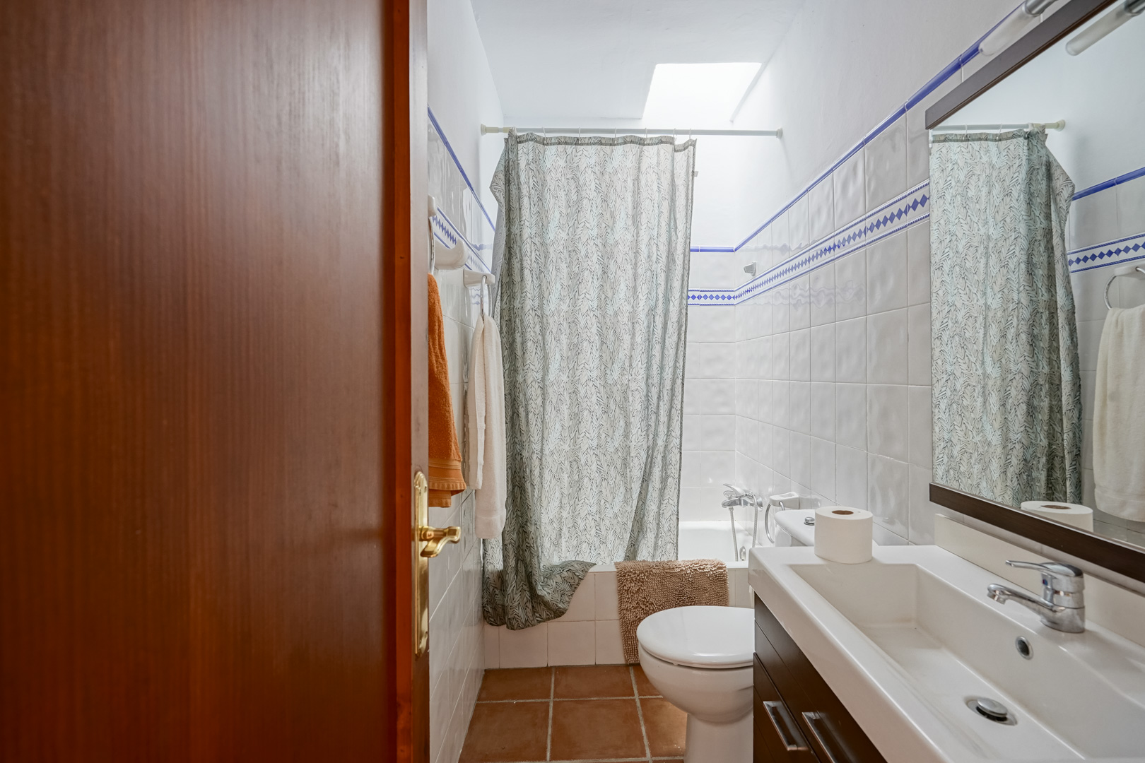 Salle de bain avec carrelage blanc à liseré bleu, meuble vasque moderne et baignoire bénéficiant d'un éclairage naturel par puits de lumière.