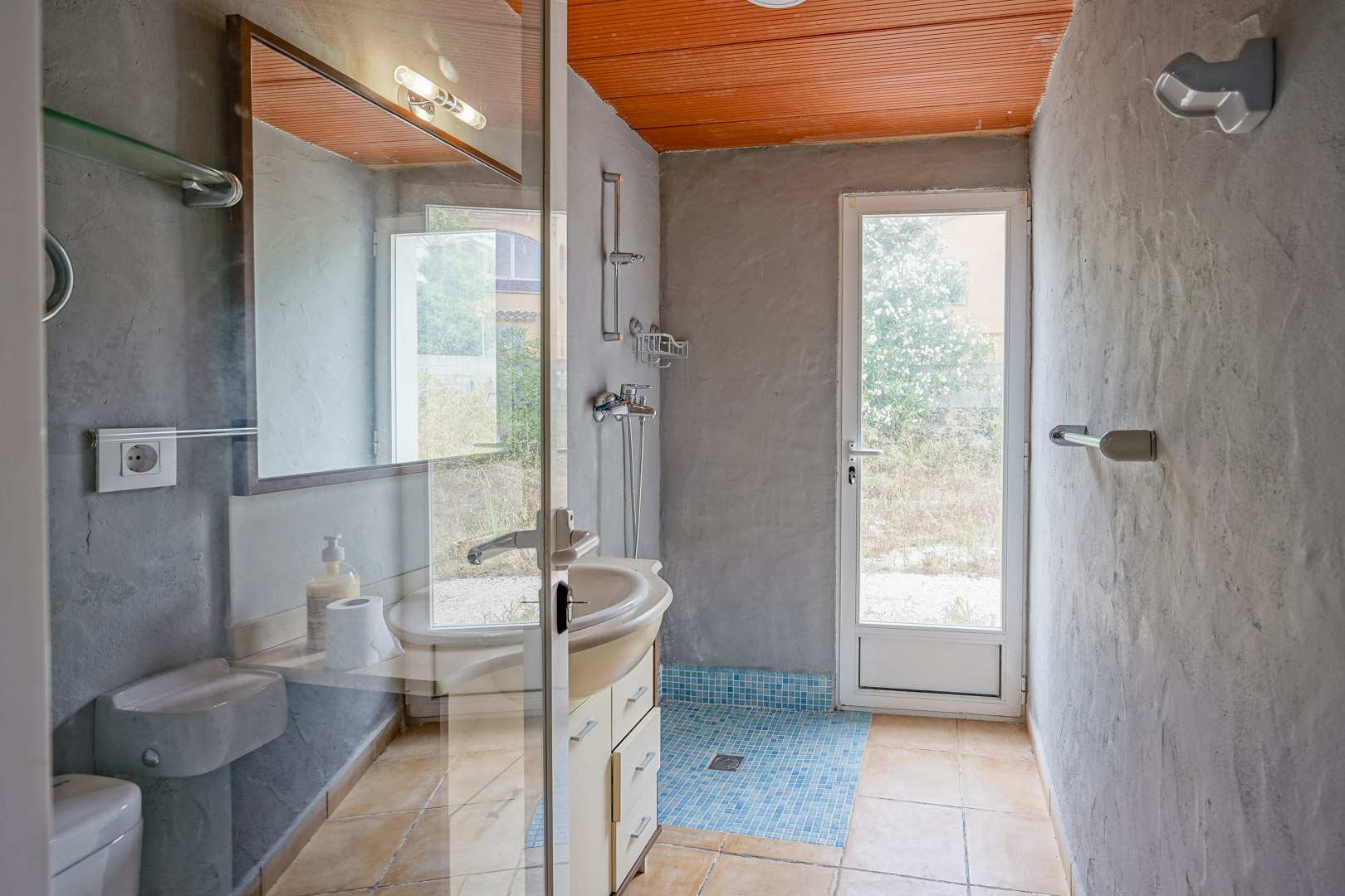Salle de bain avec murs en crépi gris, plafond à lattes orange et douche en mosaïque bleue. Une porte vitrée offre un accès direct vers l'extérieur de la propriété.