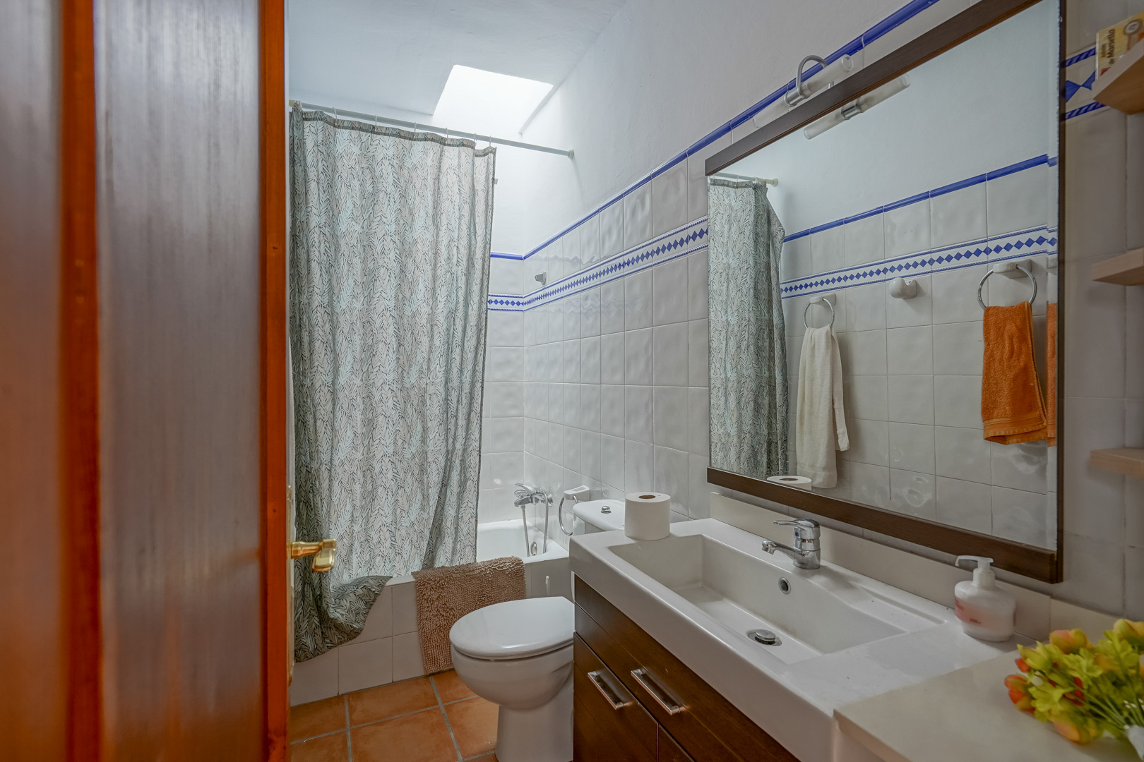 Salle de bain fonctionnelle avec carrelage blanc à liseré bleu, meuble vasque en bois sombre et baignoire éclairée par un puits de lumière naturel.