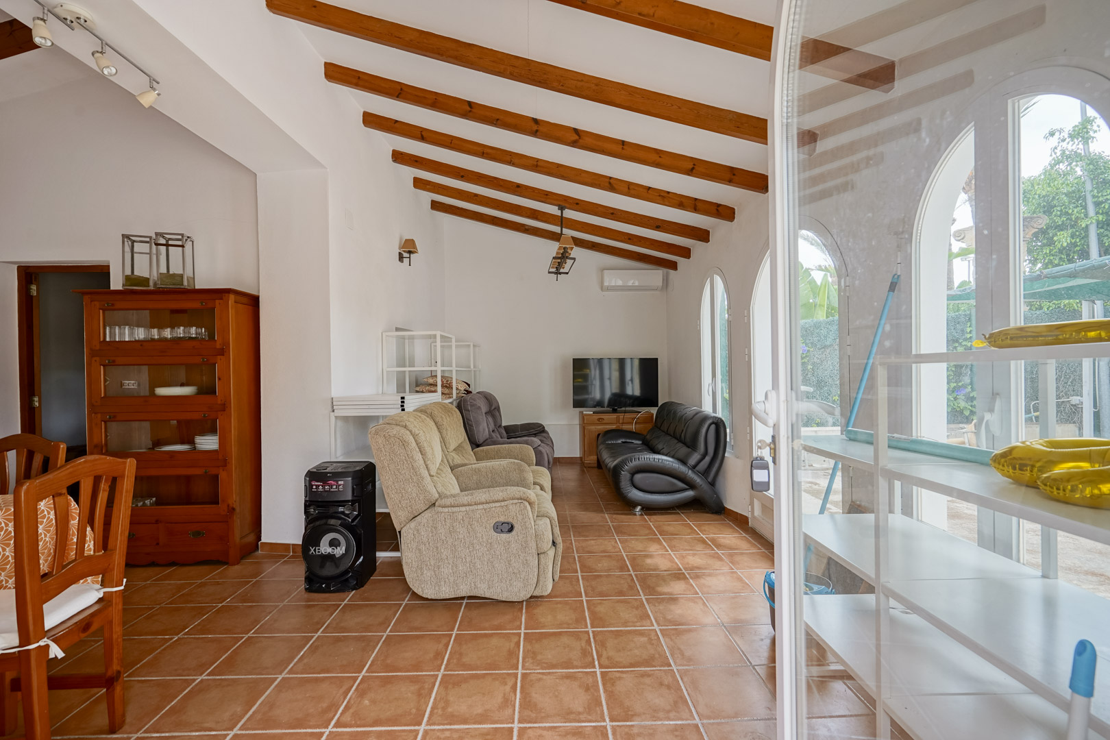Salon avec plafond incliné à poutres apparentes, sol en terre cuite et grandes fenêtres cintrées ouvrant sur l'extérieur.